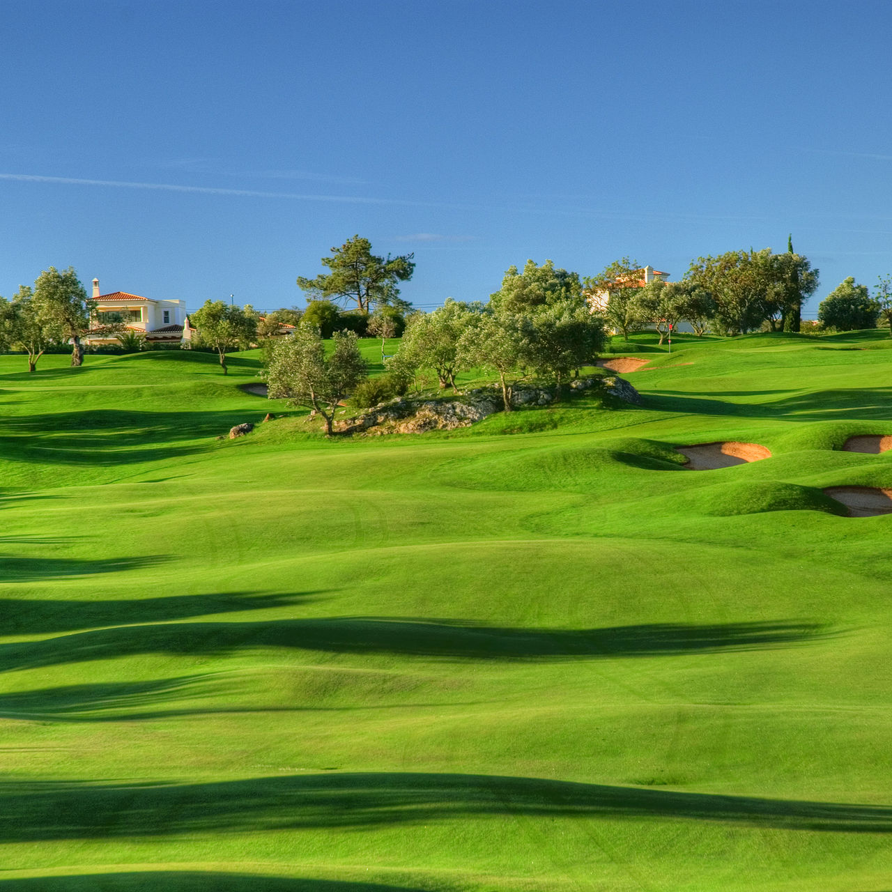 Campo de golf verde en terreno ondulado del Gramacho, con cielo azul, árboles y arbustos y algunas casas al fondo