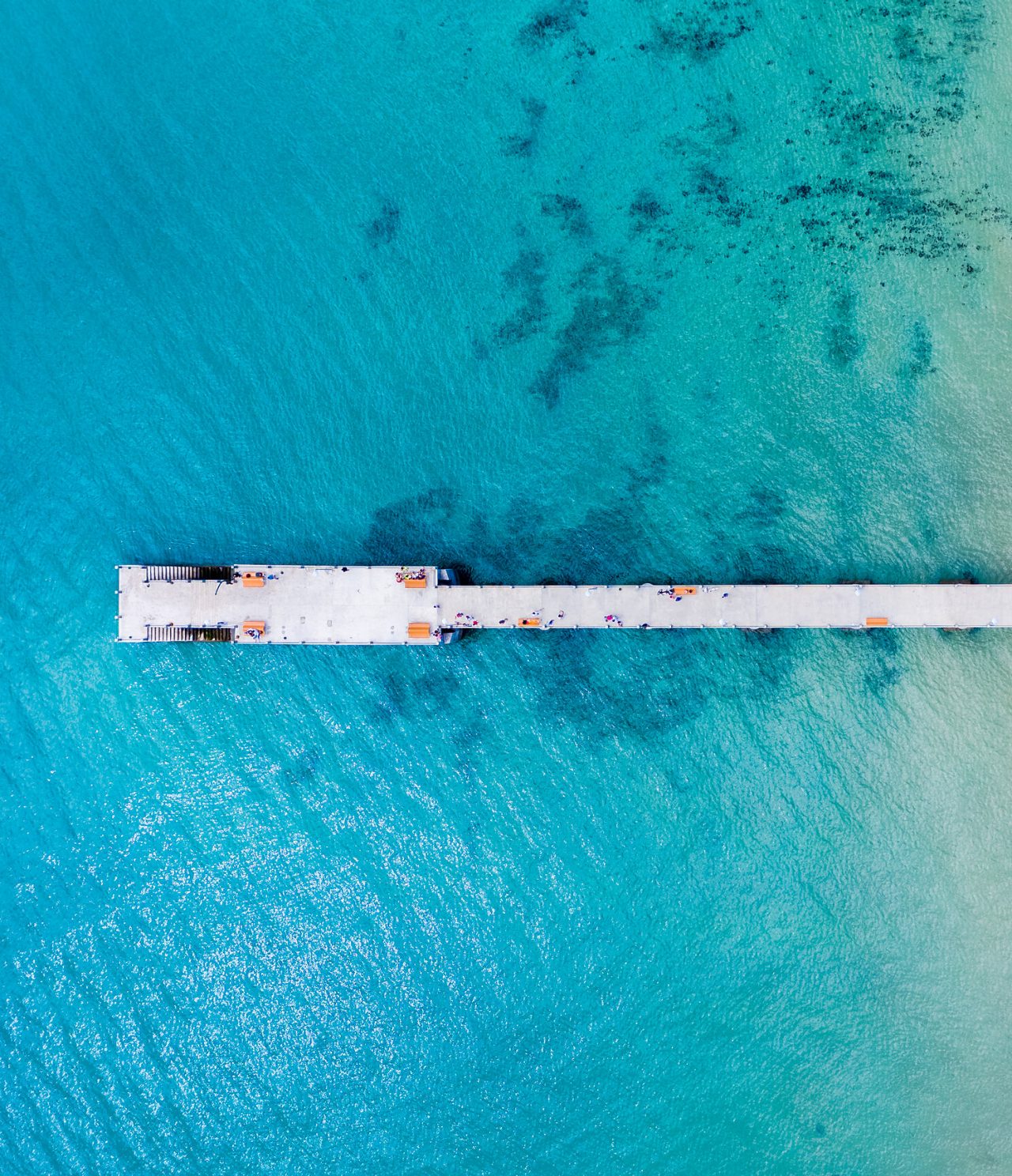 Vista aérea de un muelle de cemento, rodeado de mar azul turquesa, con escaleras para bajar al agua