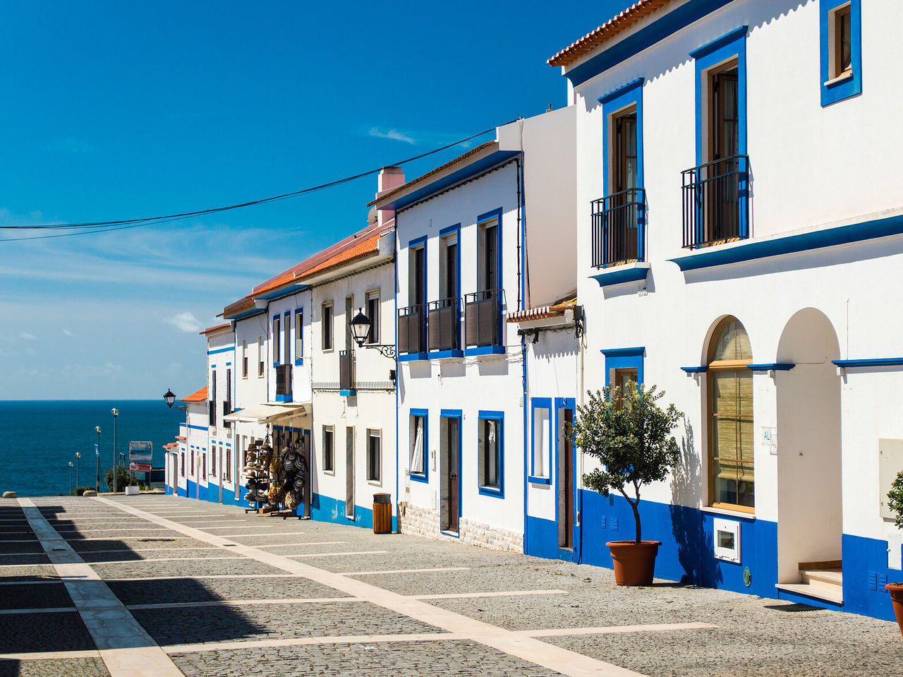 Rua com casas típicas da região do Alentejo, brancas e azuis com vasos à porta e com uma vista incrível para o oceano