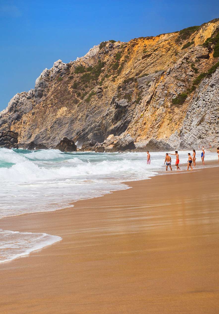 Praia em Sintra, com areia dourada e ondas a quebrar na costa, com pessoas a entrar na água e rochas ao fundo 
