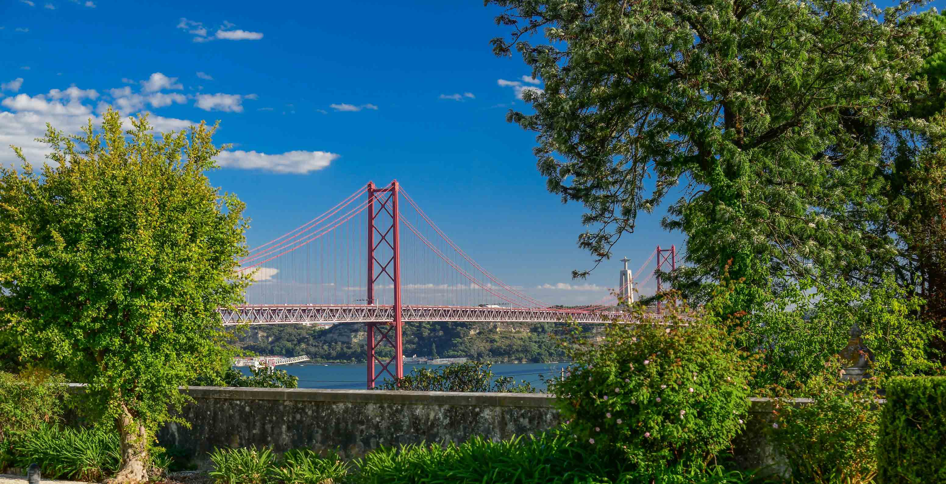 Blick vom Pestana Palace Lisboa zur 25 de Abril Brücke und dem Tejo, mit mehreren Gebäuden und Bäumen dazwischen 