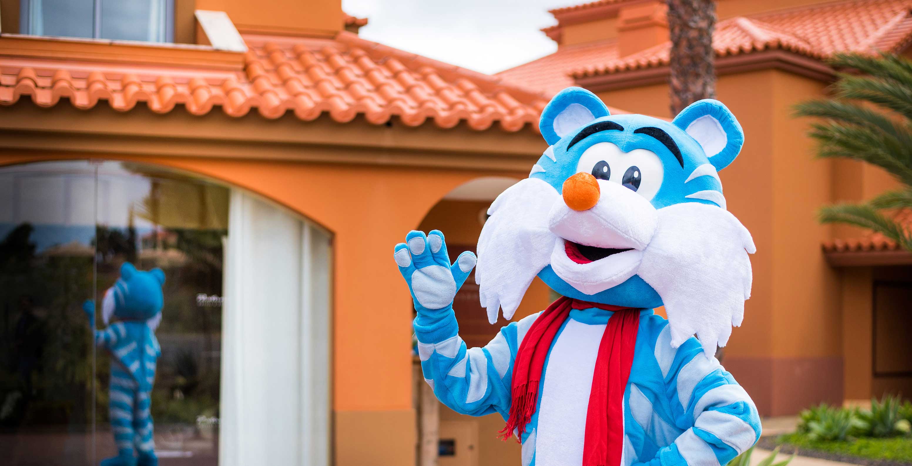 Blau-weißes Tiger-Maskottchen winkt vor dem Pestana Porto Santo, Spiegelung im Fenster sichtbar