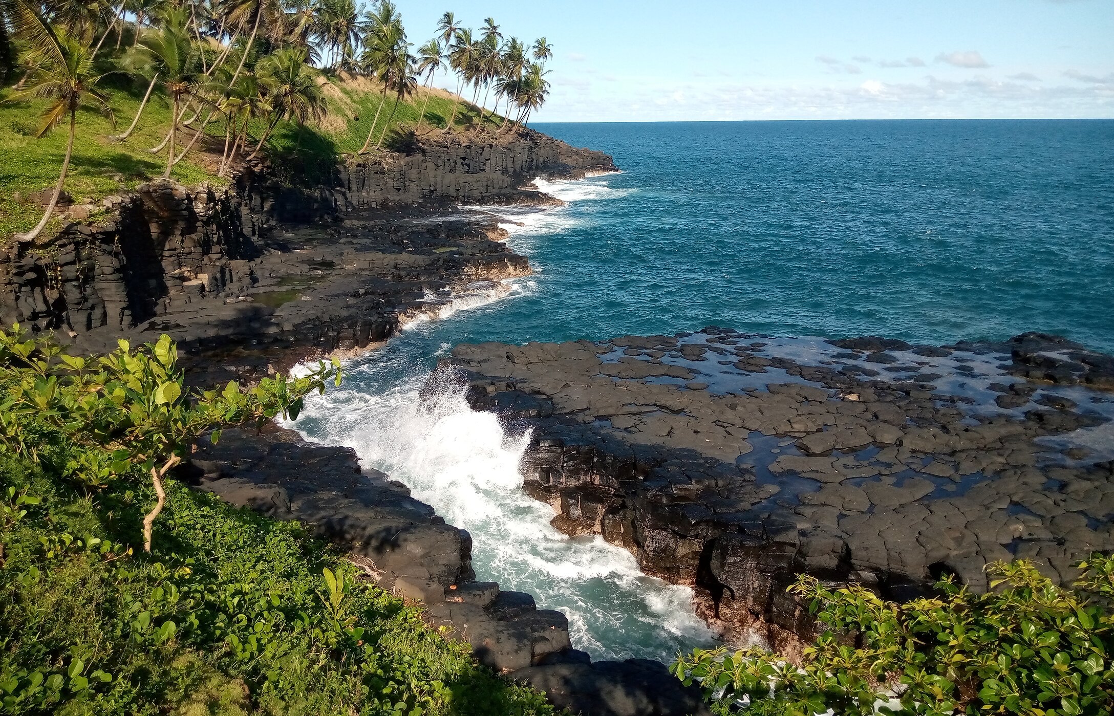 Blick auf die Hölle in São Tomé und Príncipe, mit stürmischer See, schwarzen Felsen und umliegender Vegetation.