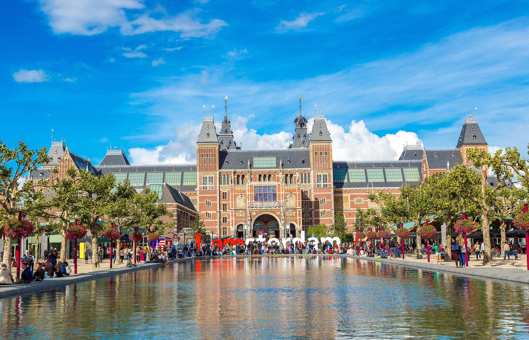 Fassade des berühmten Rijksmuseums in Amsterdam, Niederlande, mit vielen Spaziergängern, Bäumen und einem See im Vordergrund.