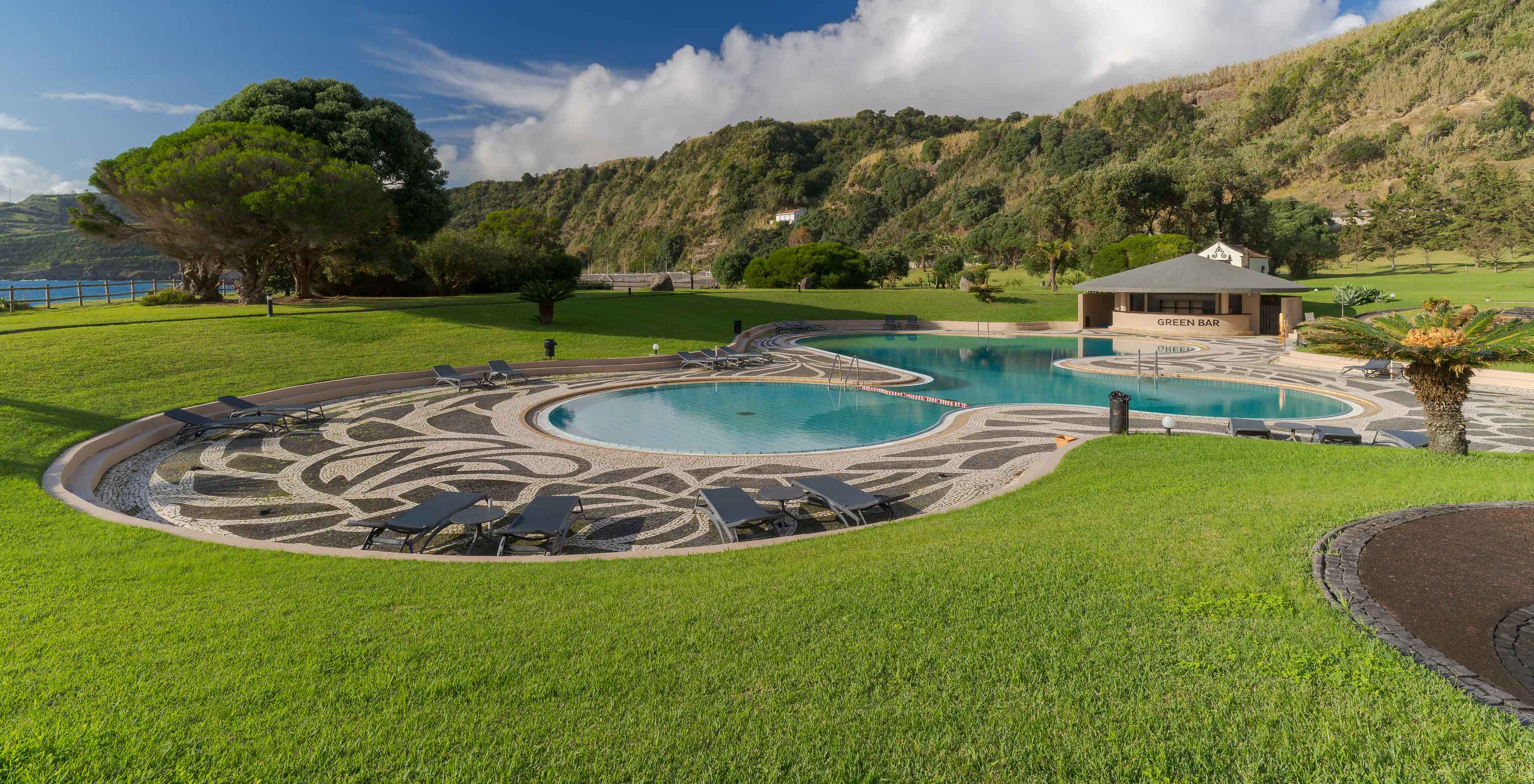 Pool at Pestana Bahia Praia surrounded by garden and hills, with bar in the back and partly cloudy sky