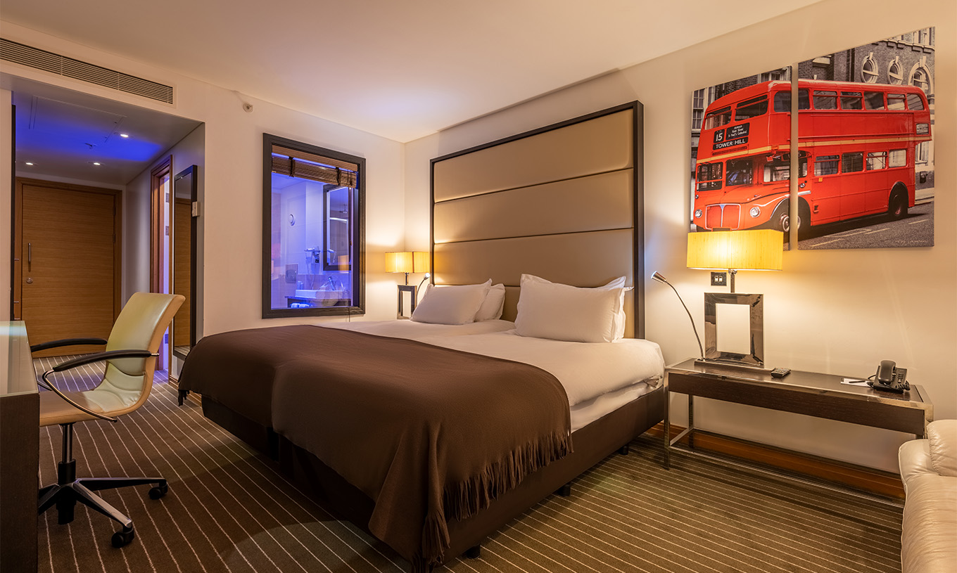 The Twin Deluxe at Pestana Chelsea Bridge has two beds with London pictures on the wall, a bedside table, sofa, and chair