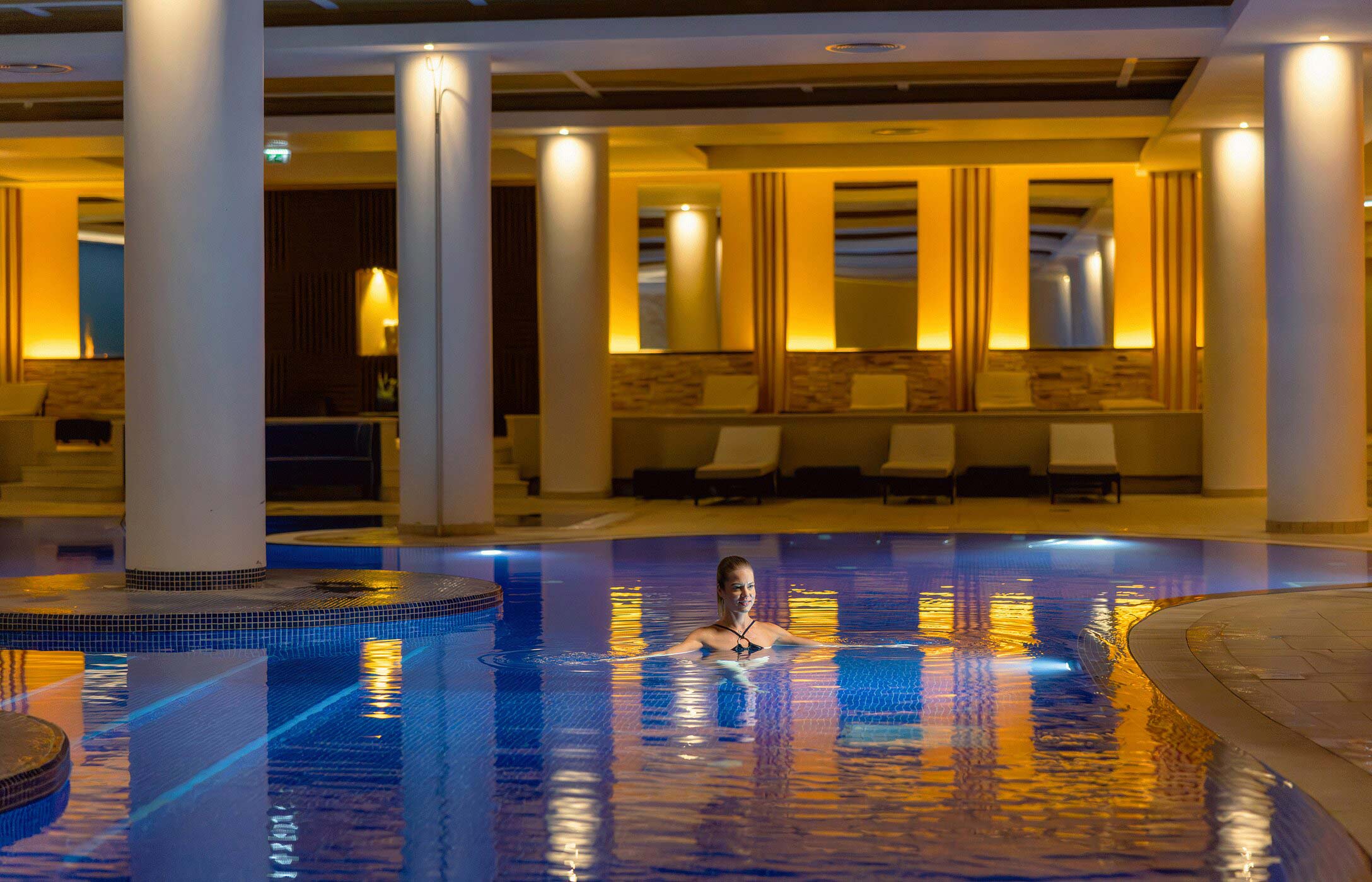 A guest swimming in the indoor pool at Pestana Casino Park Spa, illuminated by light, surrounded by columns and lounge chairs