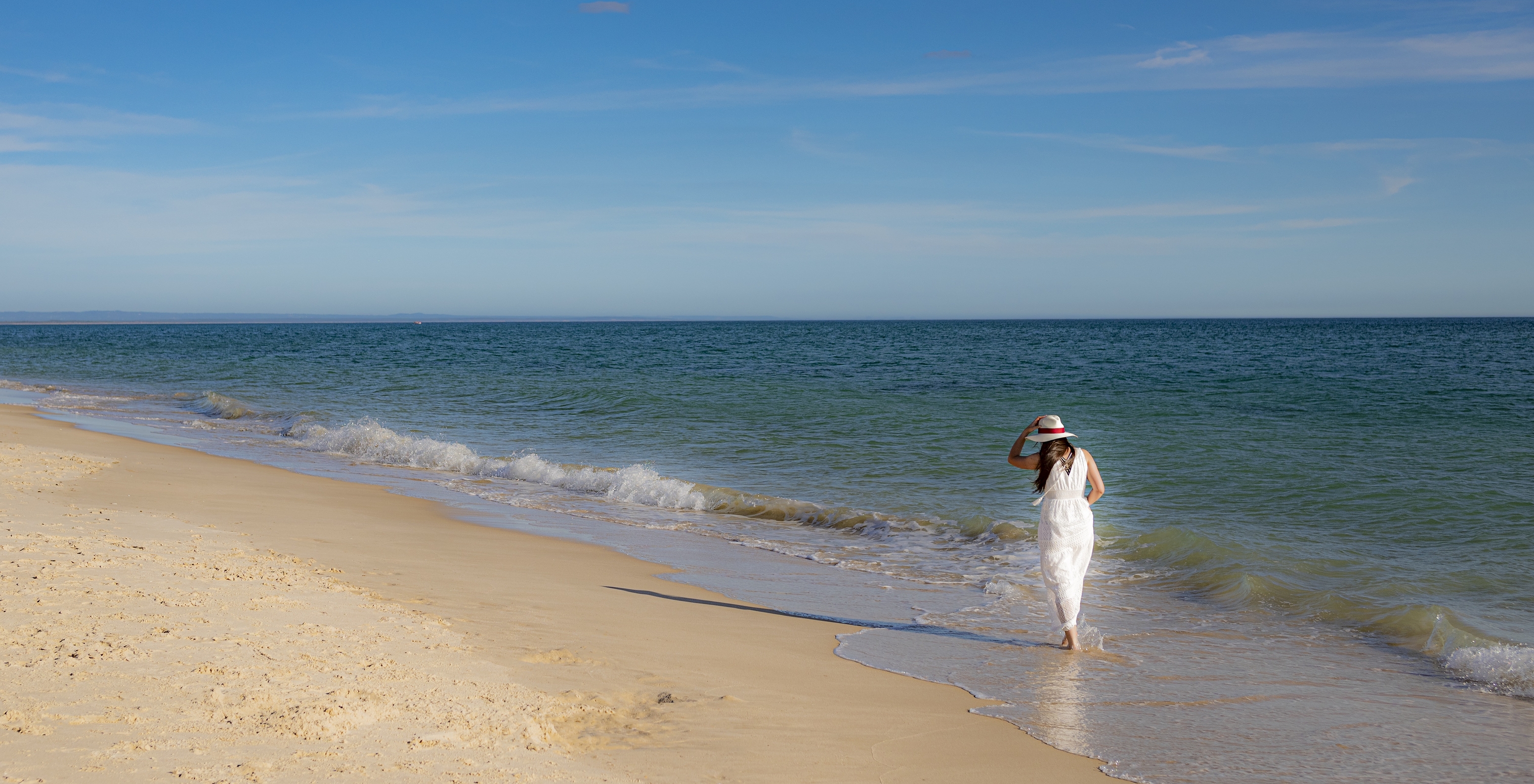 Woman in white dress and hat walks by the sea near Pestana Tróia Eco-Resort, with direct beach access