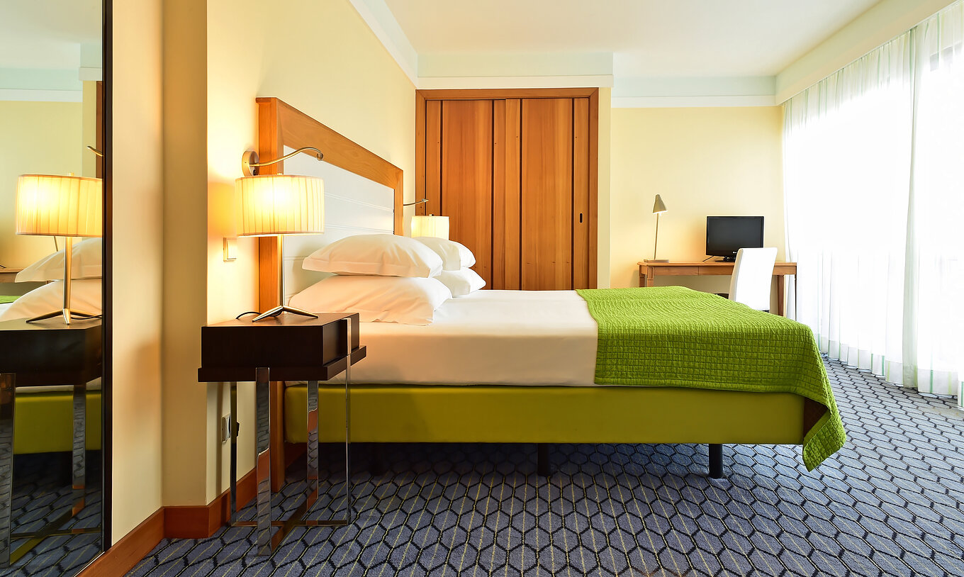 The Junior Suite of Pestana Promenade has a double bed with a green bedspread, a wardrobe, and a desk