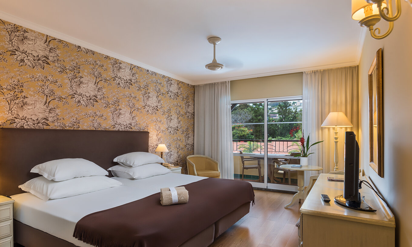 The Studio Superior at Pestana Village has a double bed with headboard and brown bedspread, floral wallpaper and balcony