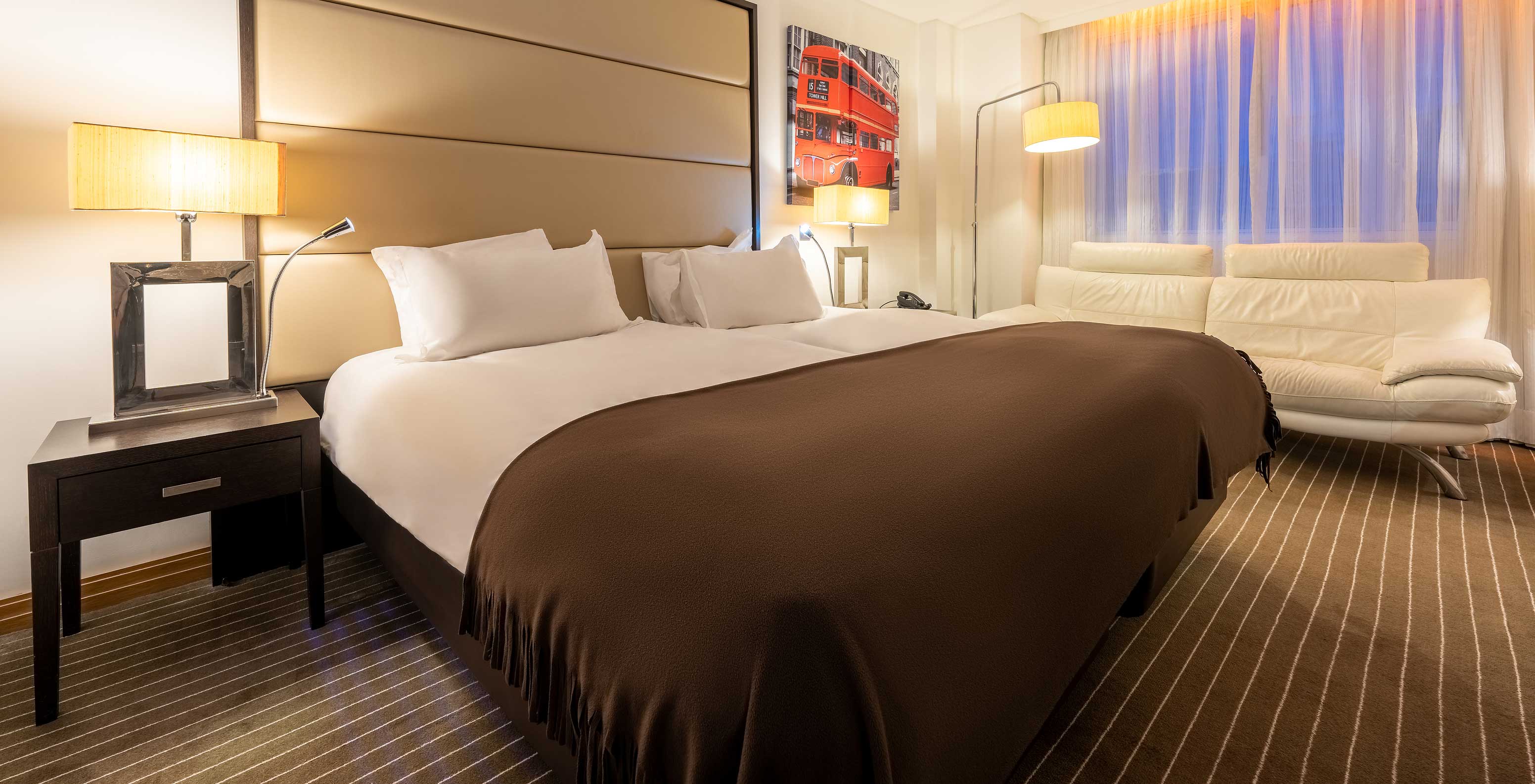 The Premium Twin at Pestana Chelsea Bridge has two beds together, nightstand, sofa, lamps and windows with curtains