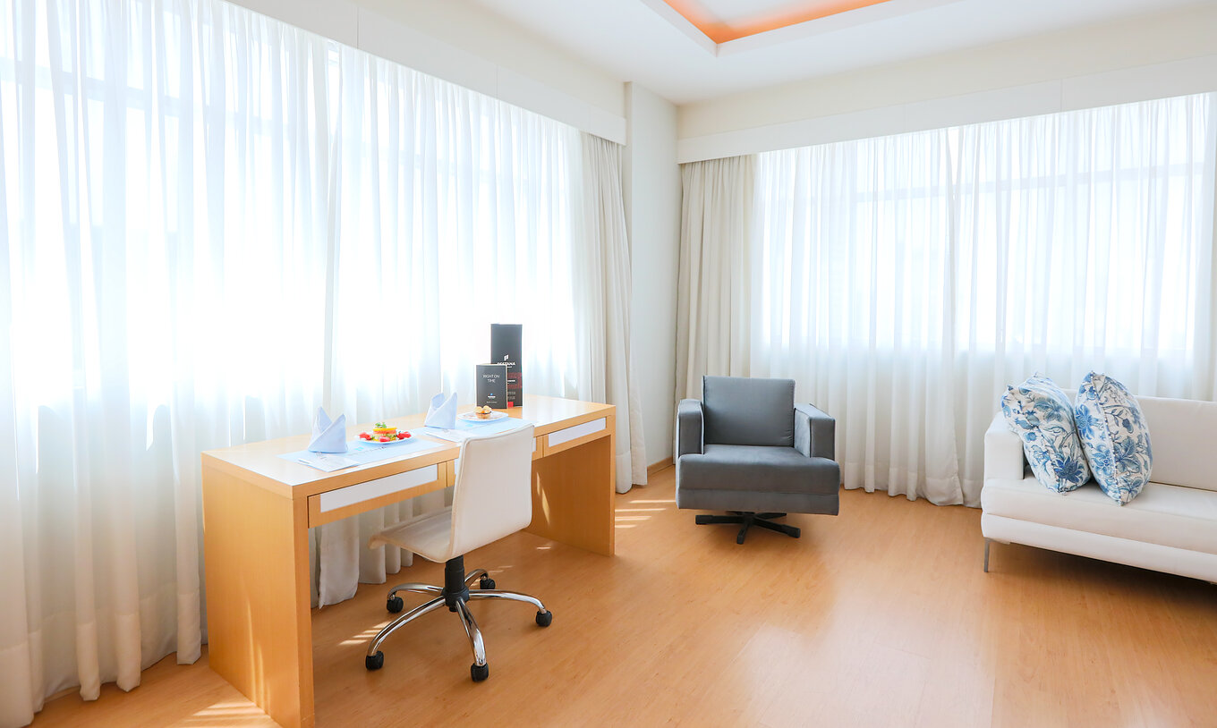 El Queen Luxo Corner del Pestana Curitiba tiene sofás y una mesa junto a dos ventanas grandes que dejan entrar bastante luz