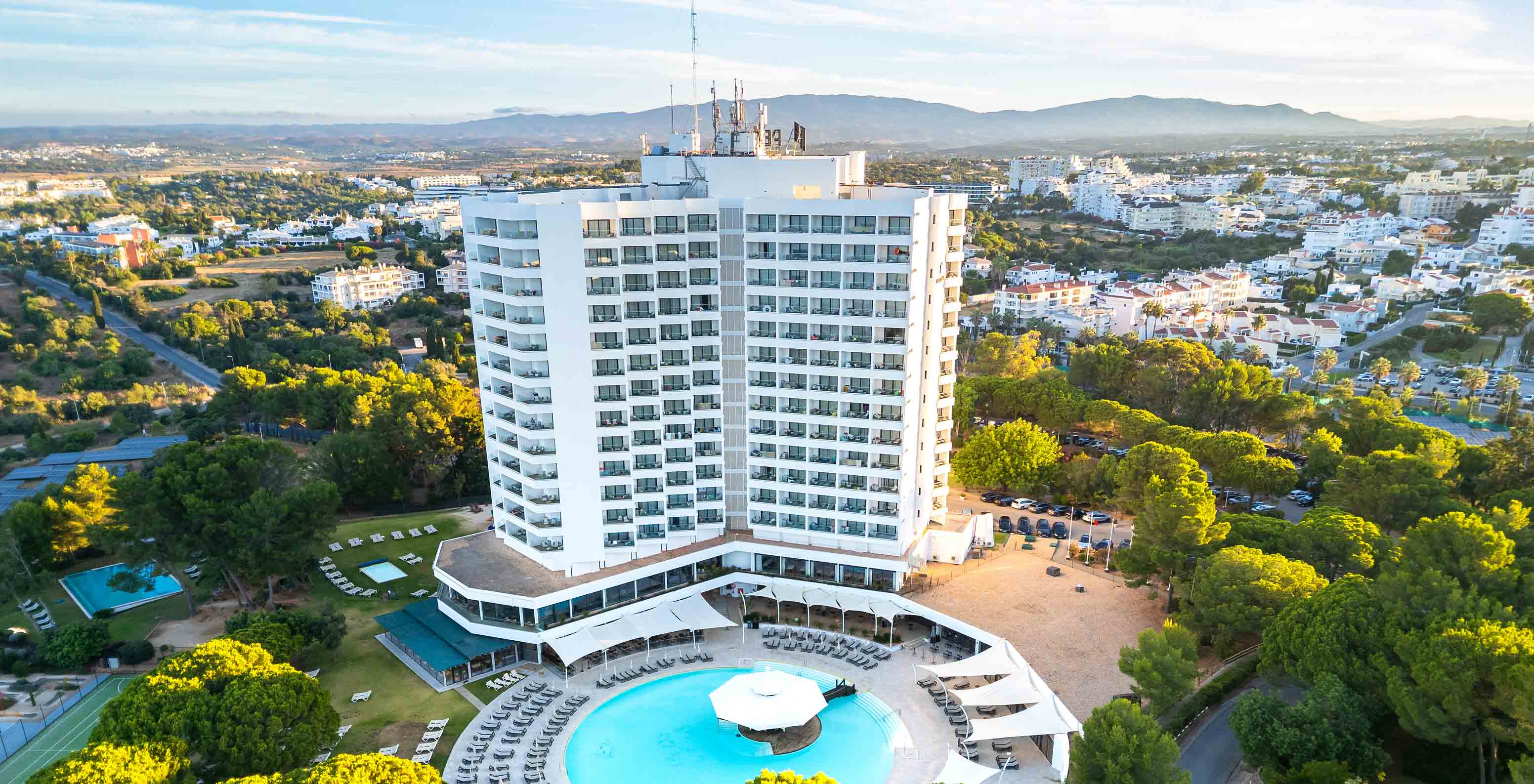 Edificio de hotel de 5 estrellas con piscina y cerca de la playa, rodeado de vegetación y varias sombrillas