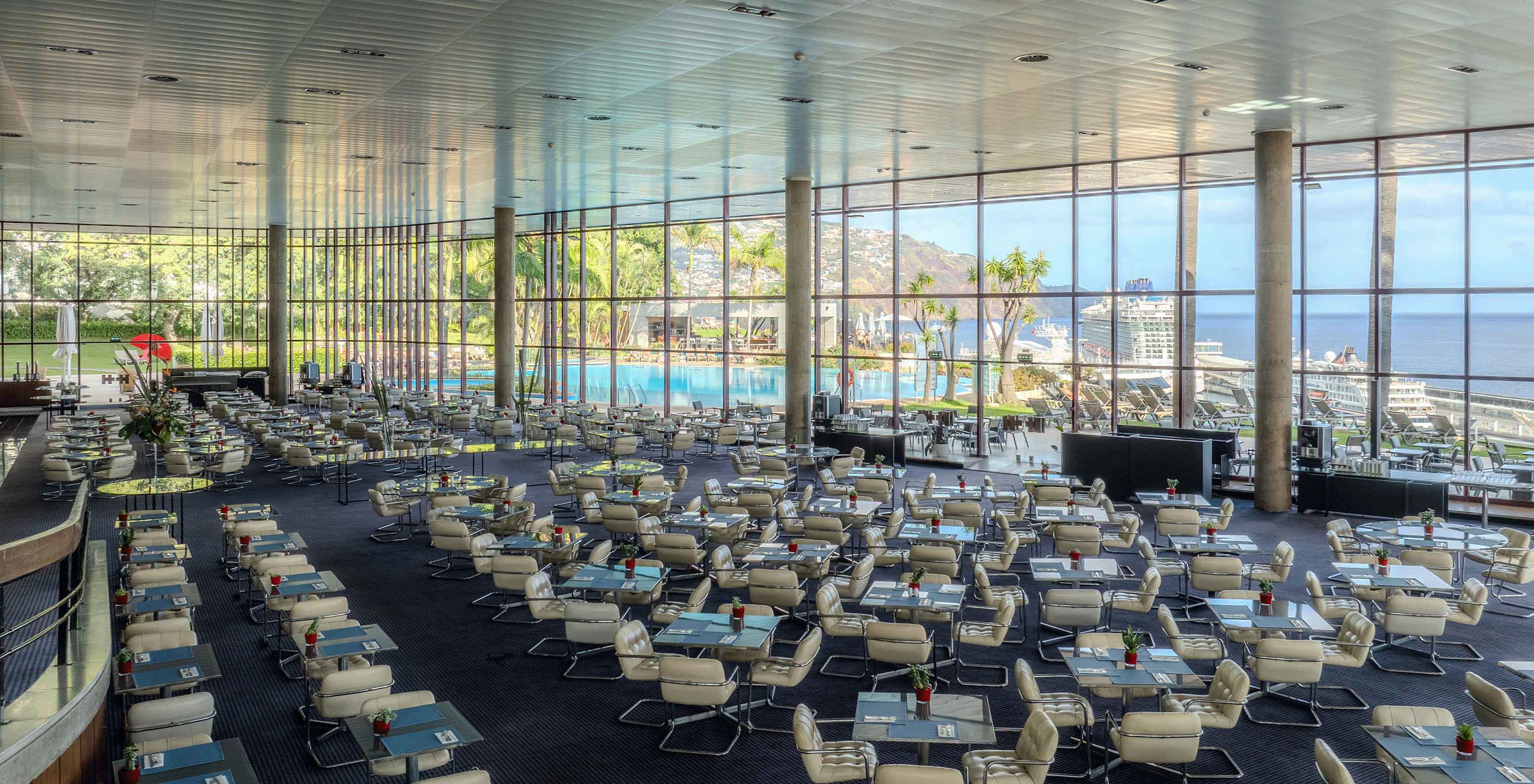 Vista panorámica de un restaurante con varias mesas, una rampa y un gran cristal con vistas a la piscina y al mar