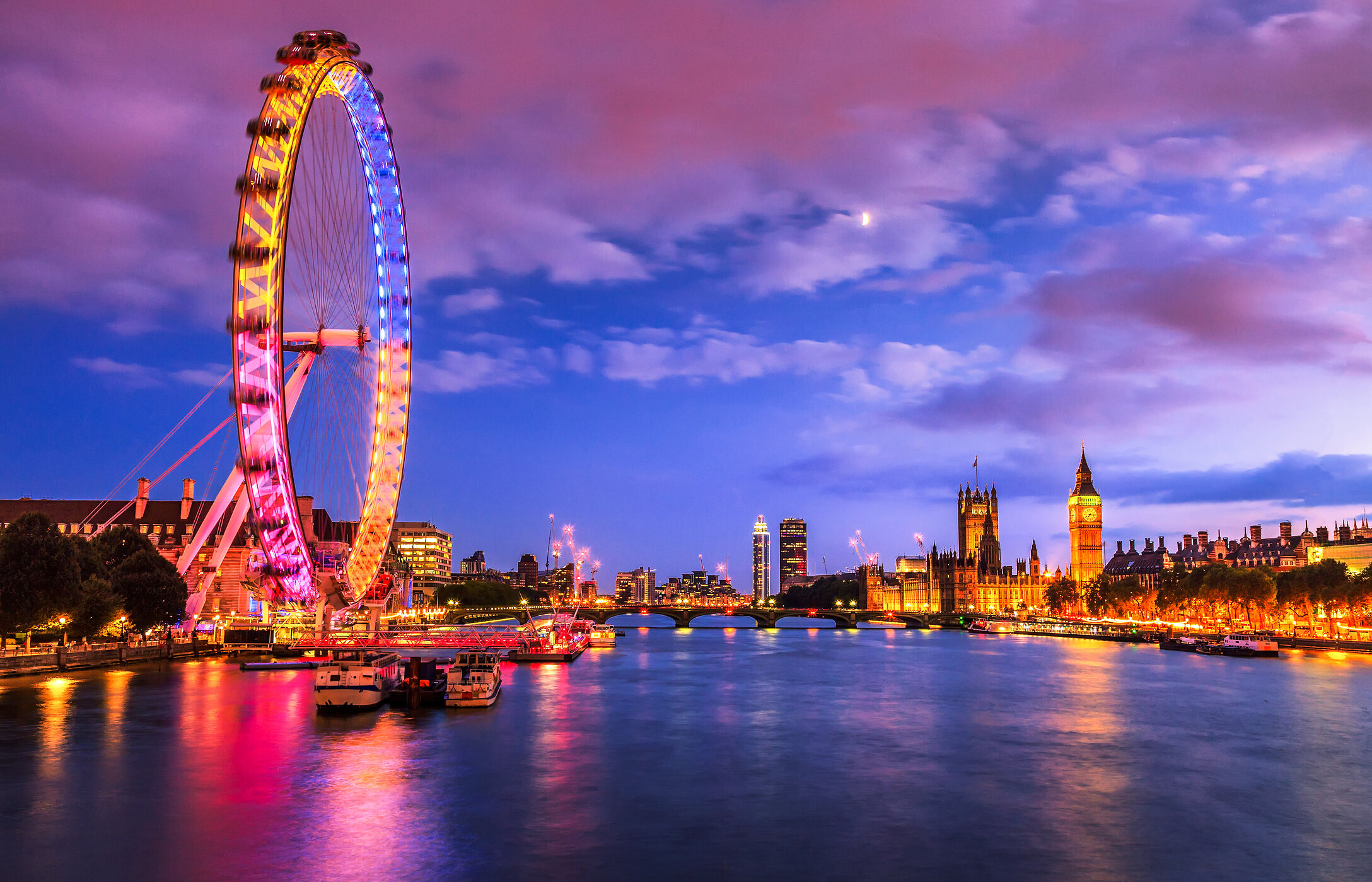 La noria London Eye en Londres, iluminada con colores vibrantes junto al río, con el Big Ben al fondo