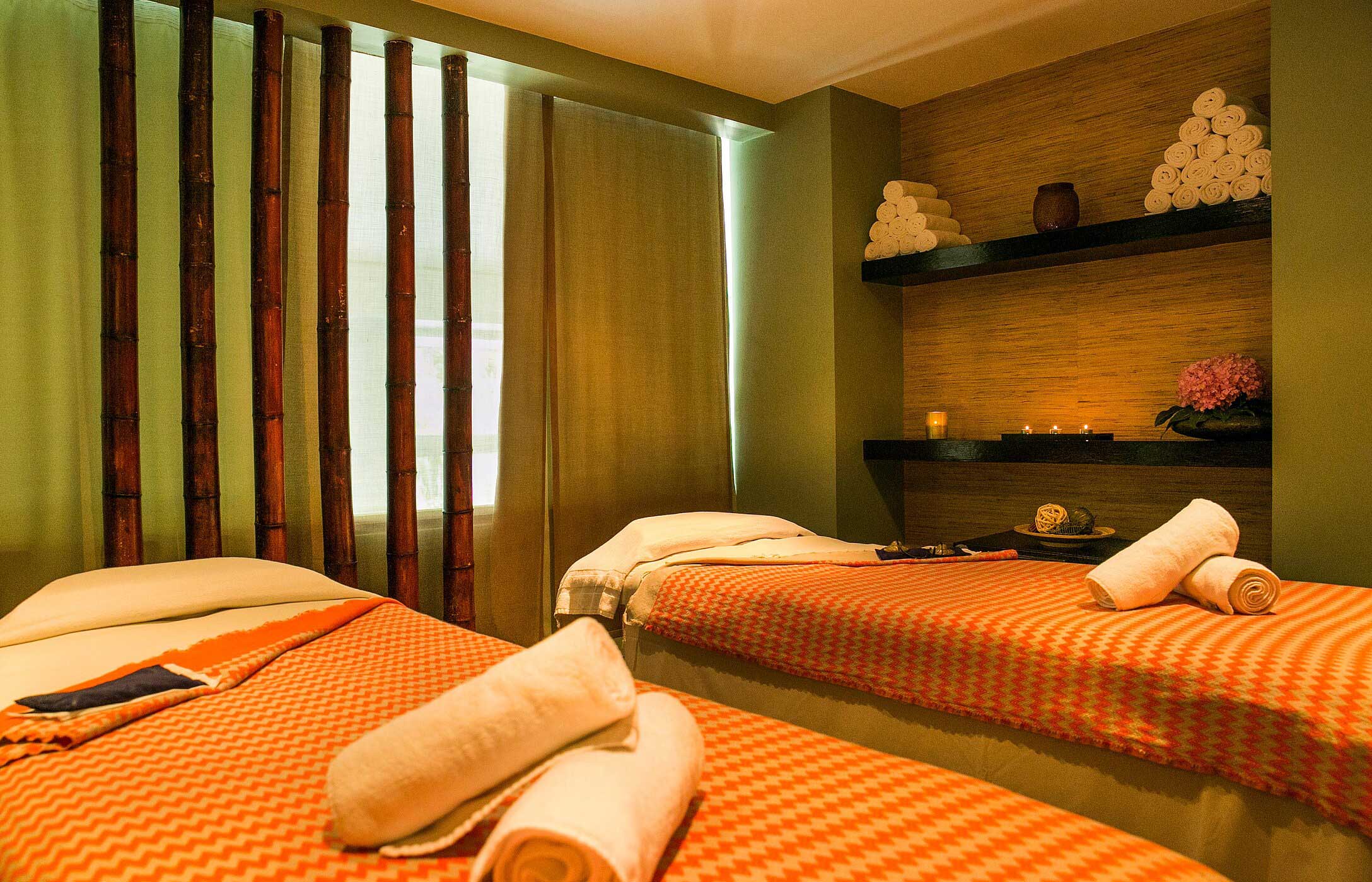 Sala de spa relajante con dos camillas de masaje cubiertas con mantas naranjas, rodeadas de bambú, en Pestana Palace Spa