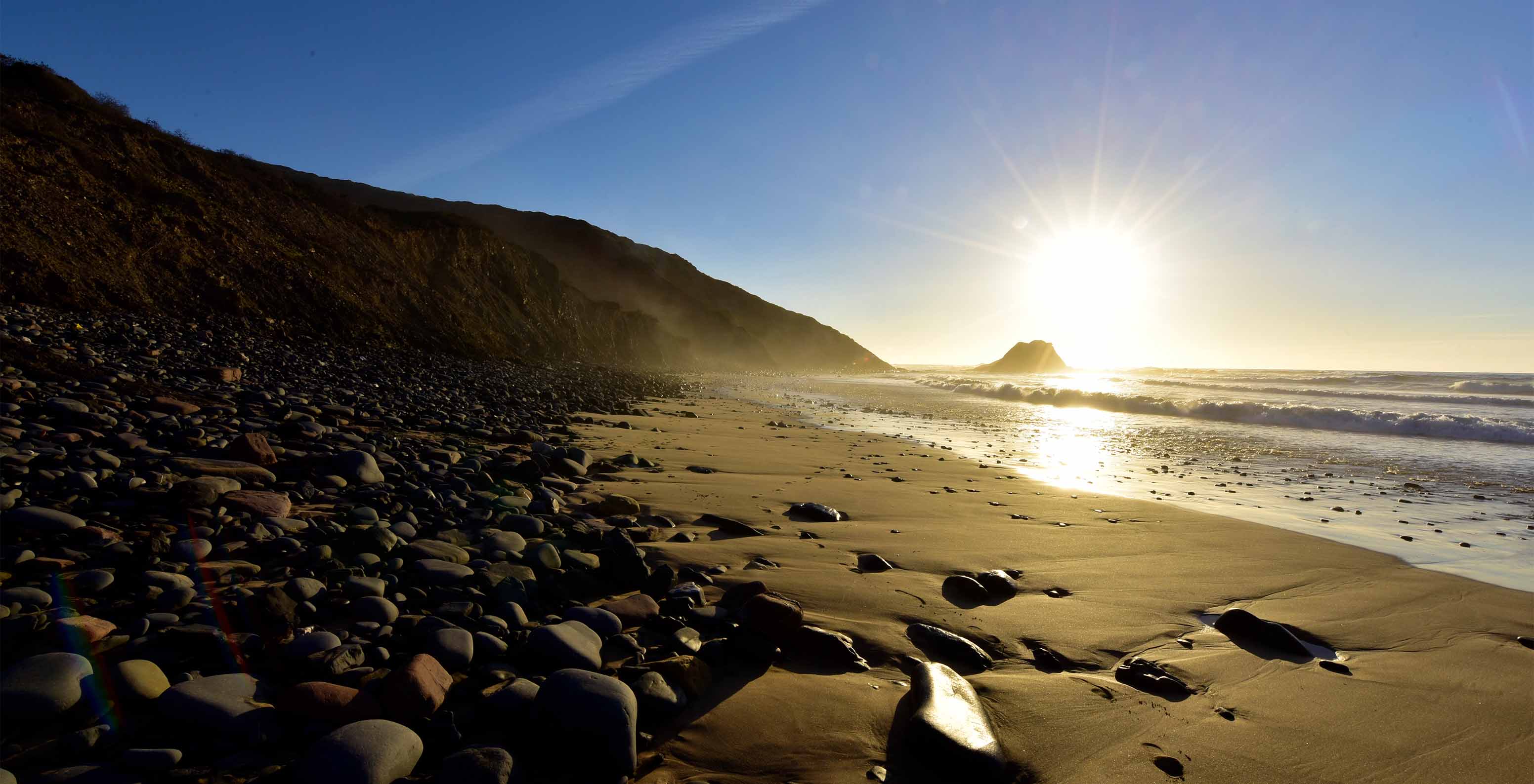 Plage au lever du soleil, avec la lumière se reflétant sur la mer, des pierres sur le sable et une falaise derrière