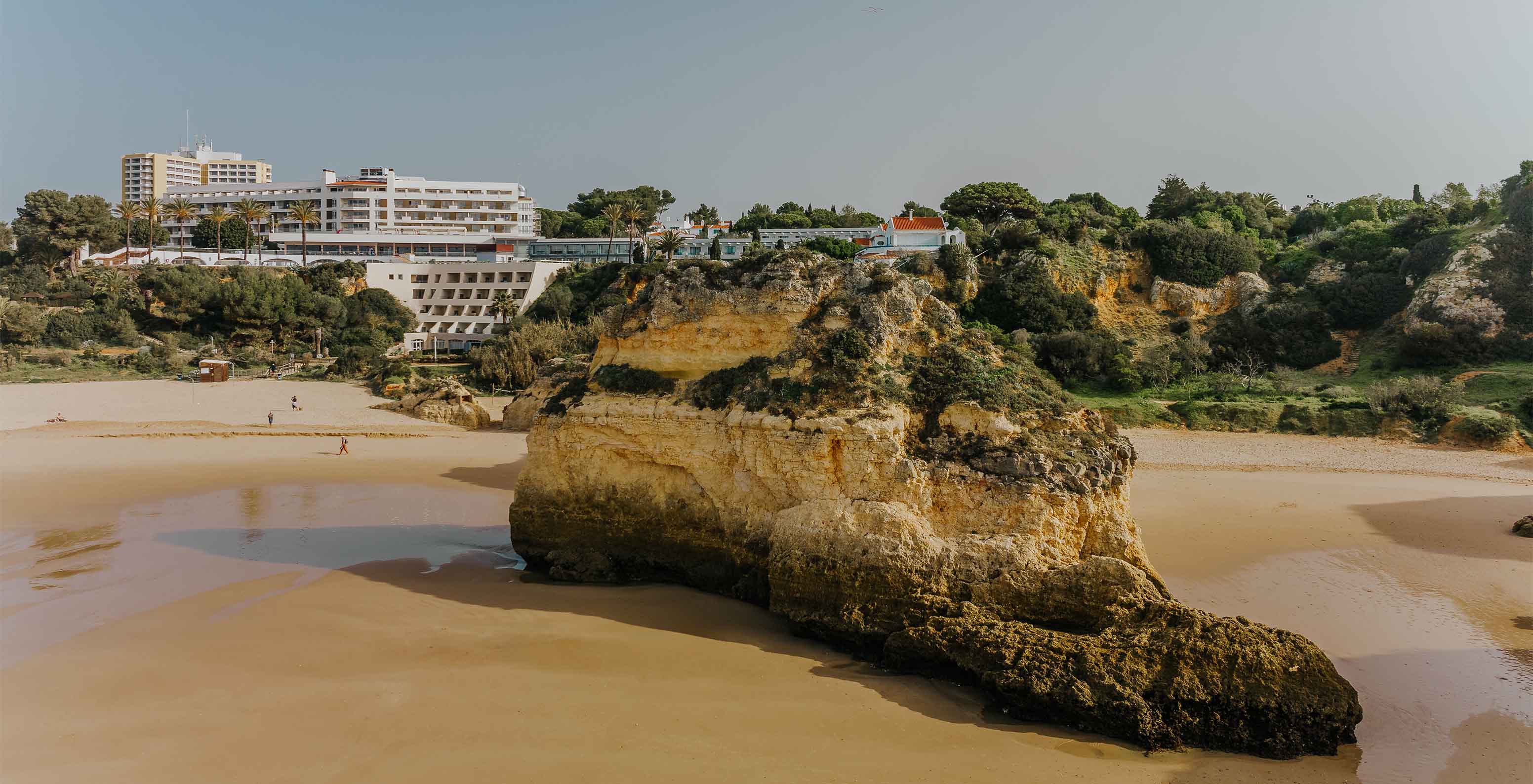 Plage d'Alvor avec rochers, sable, végétation, la falaise et des bâtiments en arrière-plan, ciel dégagé