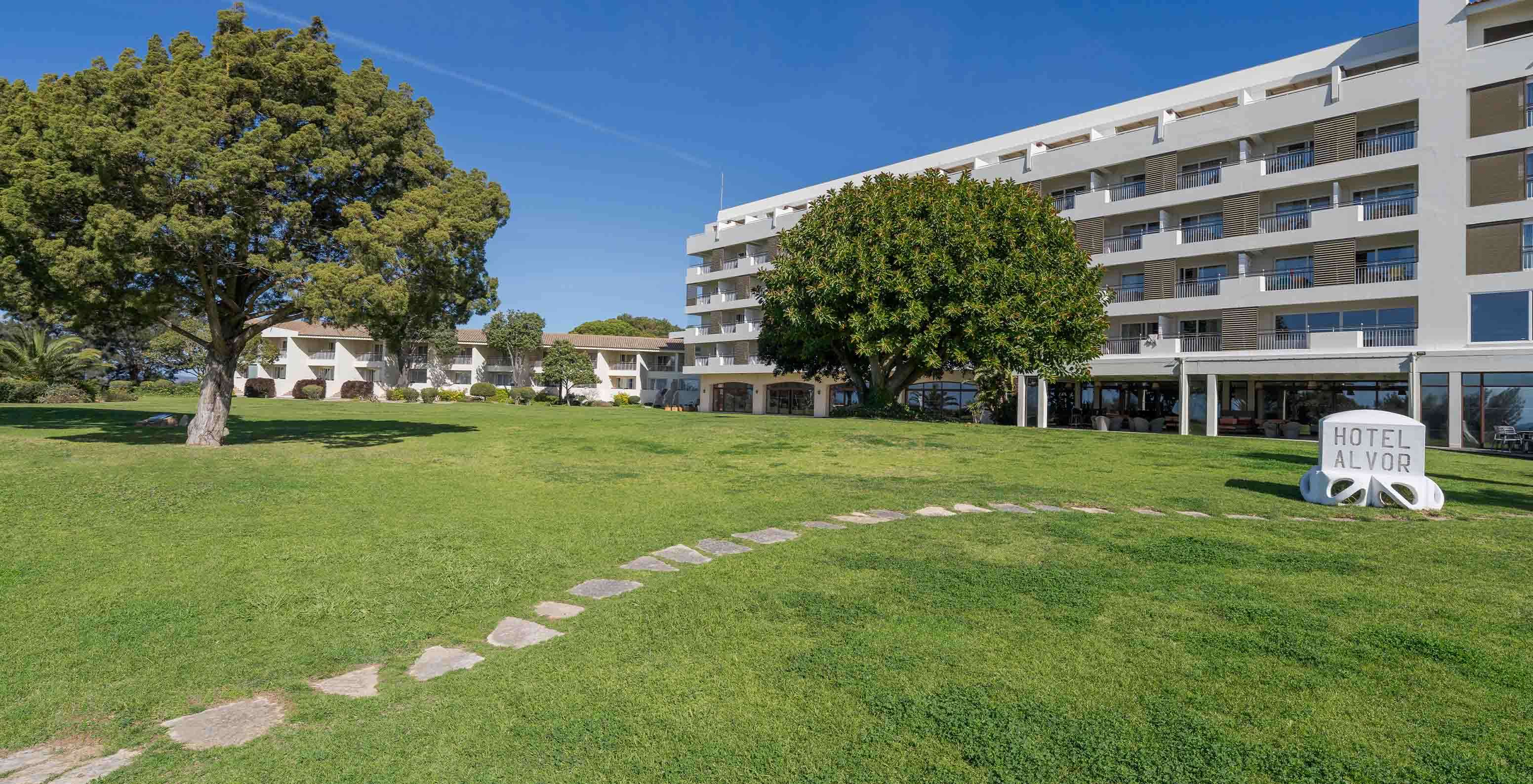 Jardin avec des arbres du Pestana Alvor Praia avec les bâtiments de l'hôtel derrière et un chemin en pierres