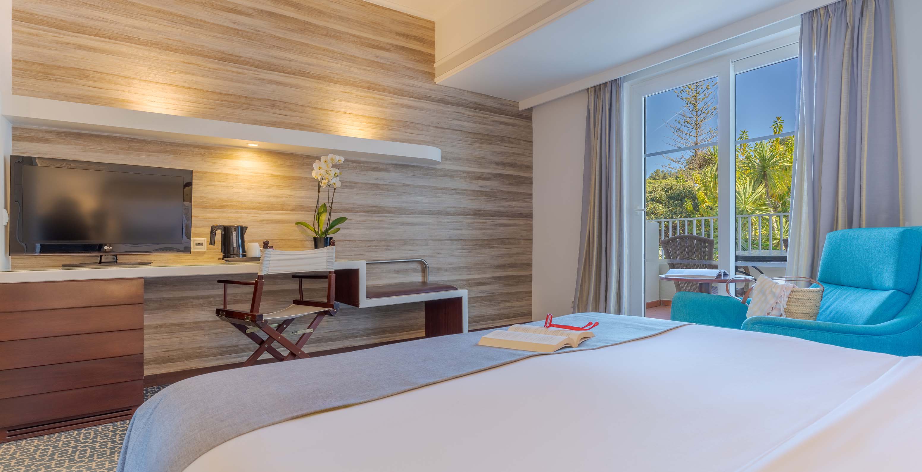 Le Deluxe du Pestana Alvor Praia a un lit double, télévision sur le bureau, fauteuil bleu et fenêtre vers le balcon