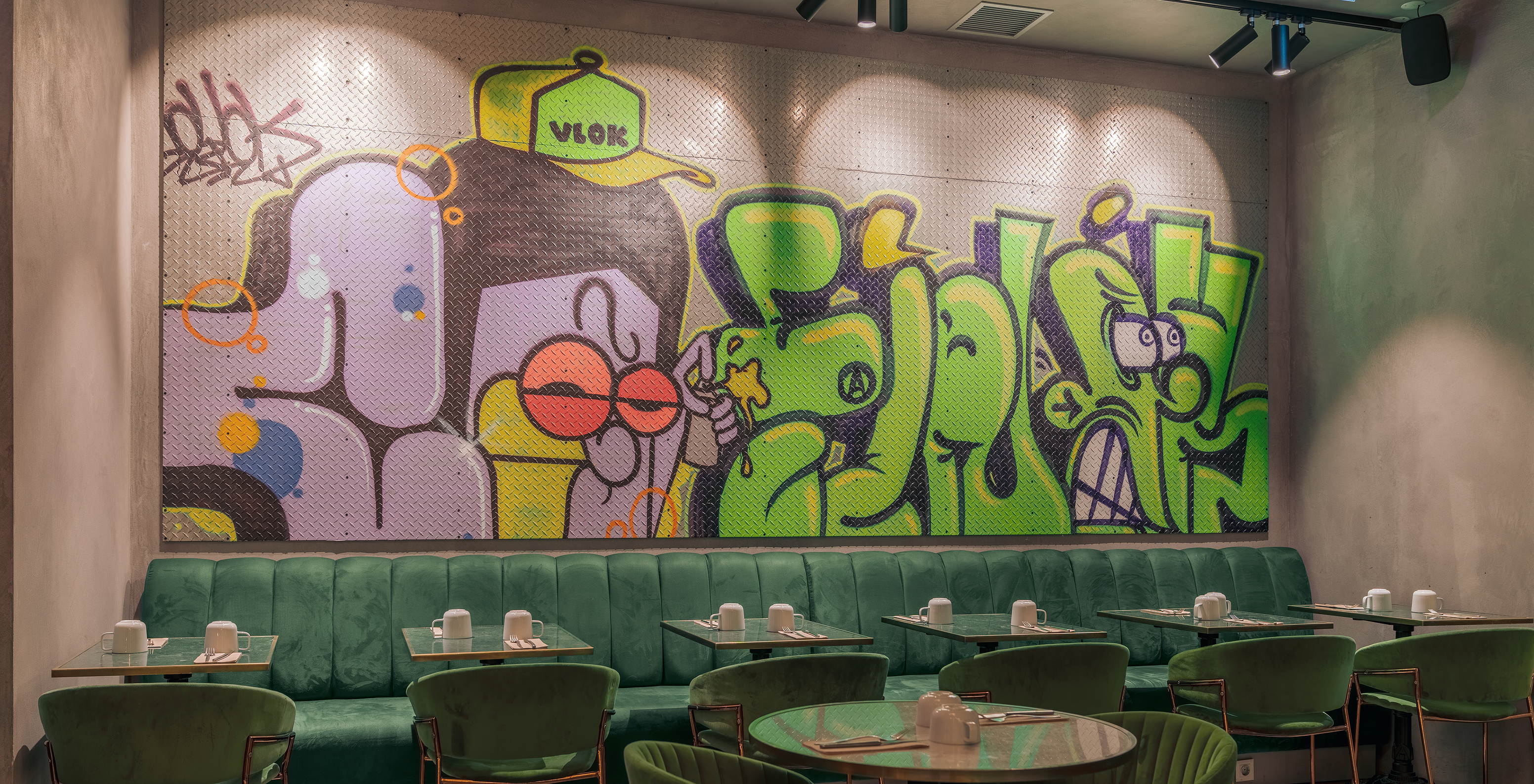 Restaurant avec tables chaises et canapés verts avec verres en porcelaine blanche et un mur coloré avec des graffitis