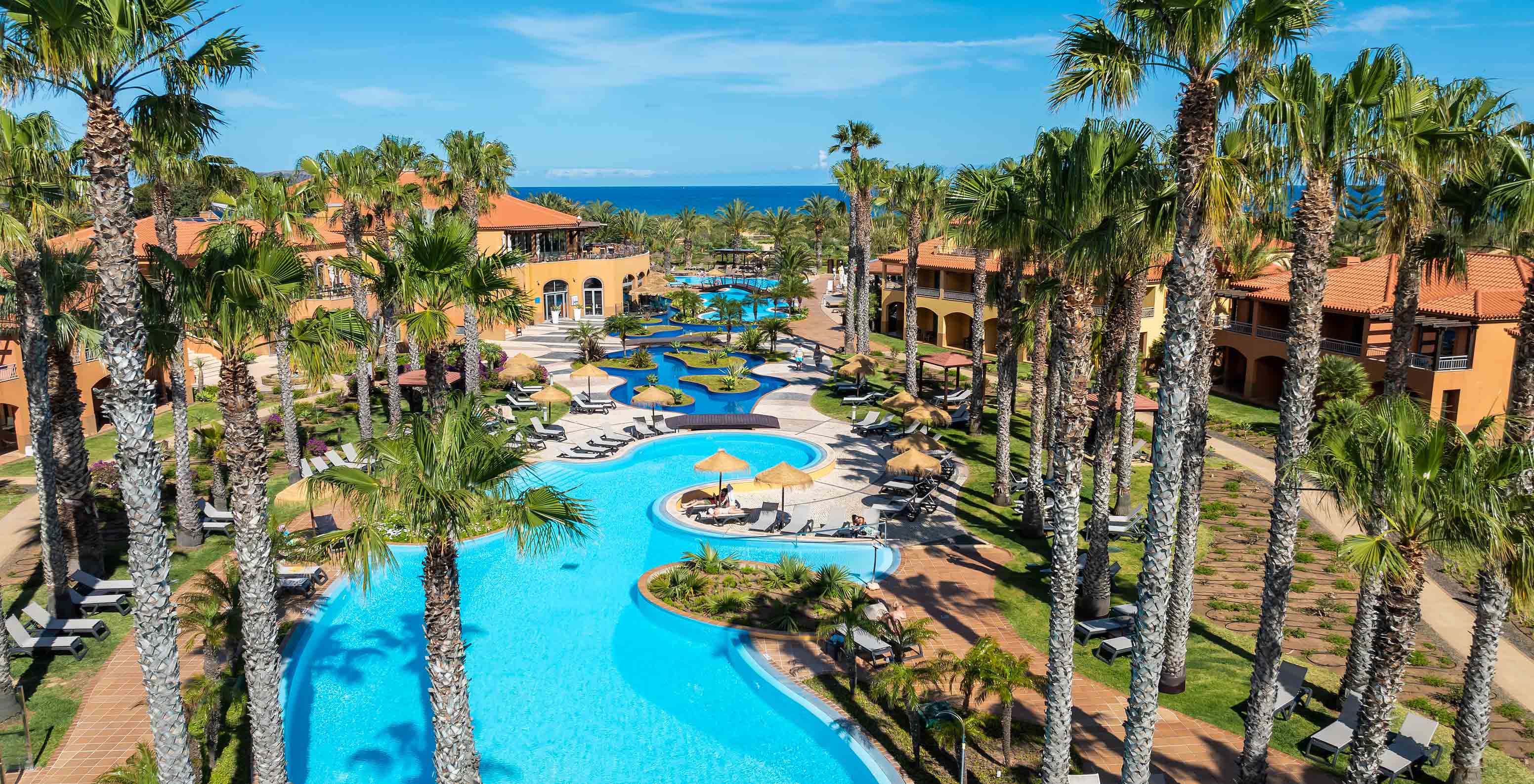 Vue de la piscine du Pestana Porto Santo entourée de palmiers et de bâtiments colorés sous un ciel bleu