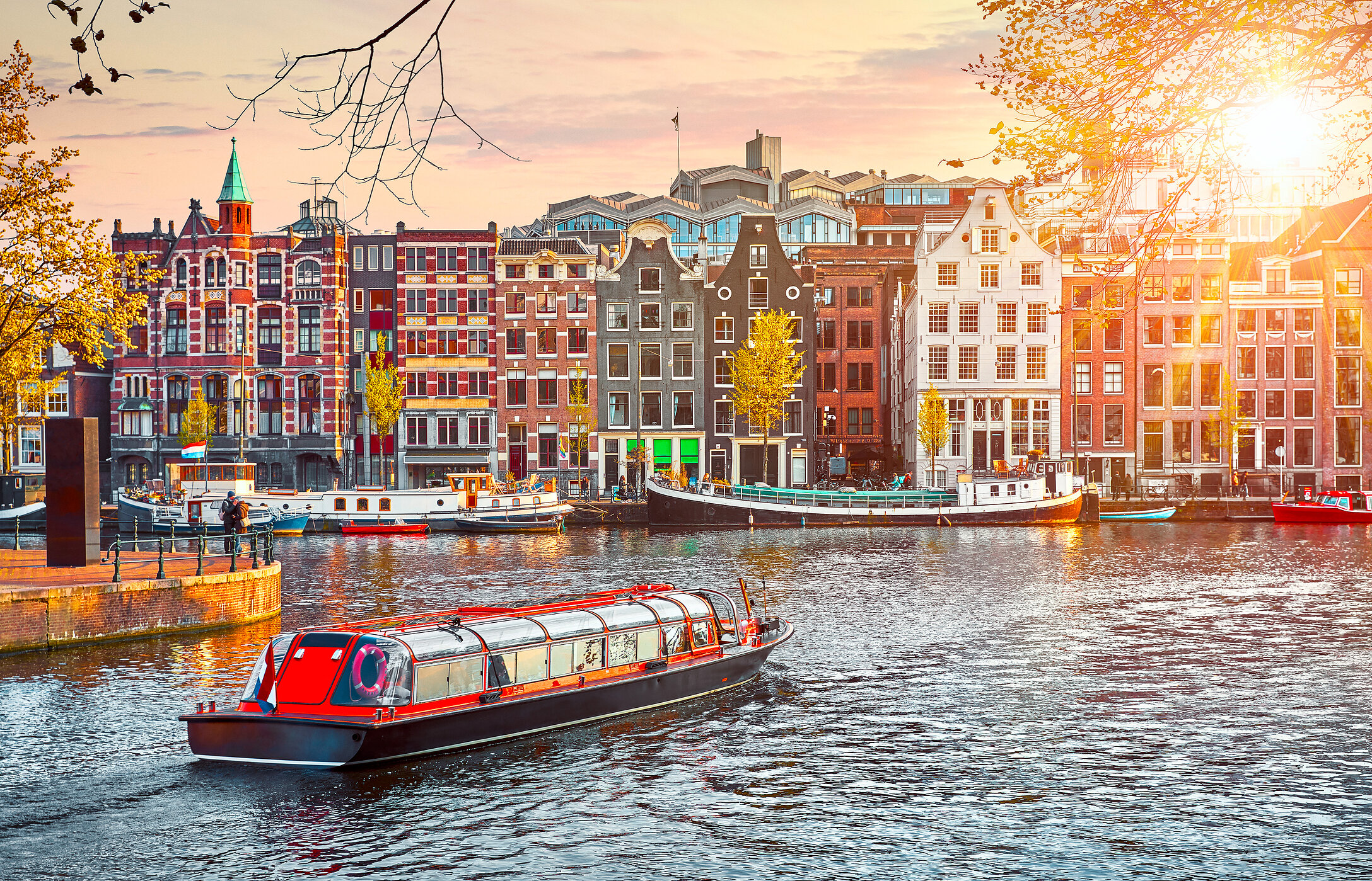 Vue pittoresque des canaux d'Amsterdam, avec des maisons typiques hollandaises, un bateau touristique rouge et un ciel aux tons chauds