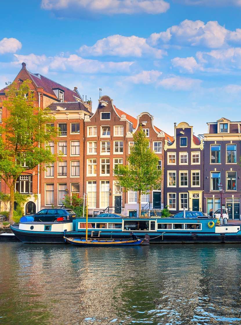 Amsterdamse gracht met mensen die langs de oever lopen, boten en typische gebouwen onder een blauwe lucht
