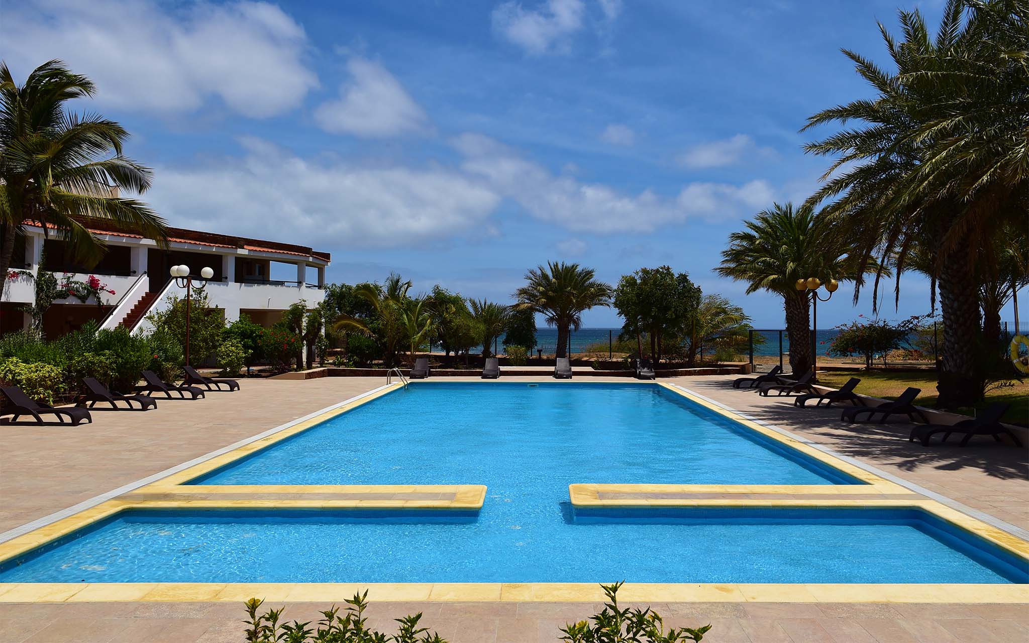 Vista da piscina exterior do Pestana Tropico, com palmeiras e espreguiçadeiras à volta da piscina, e o mar ao fundo