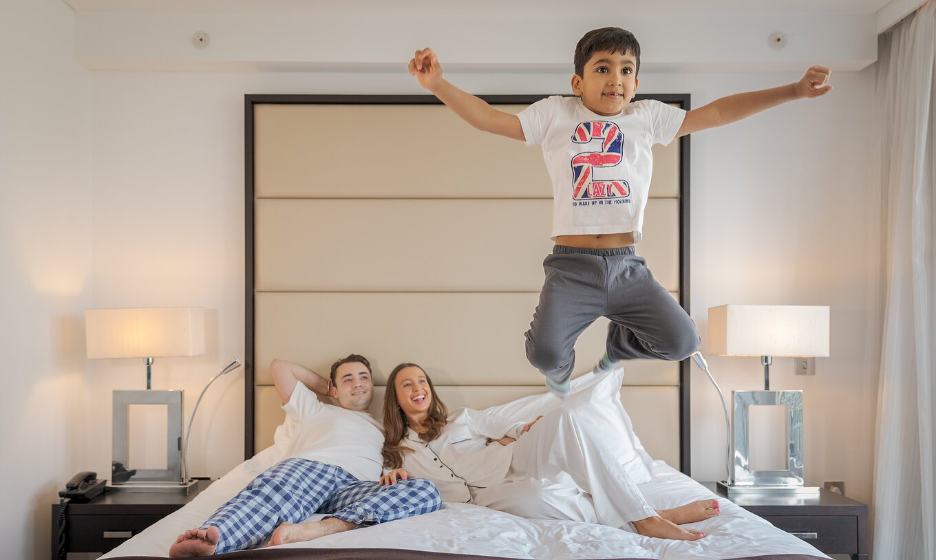 O Deluxe Family do Pestana Chelsea Bridge tem cama de casal com uma família deitada e uma criança a saltar