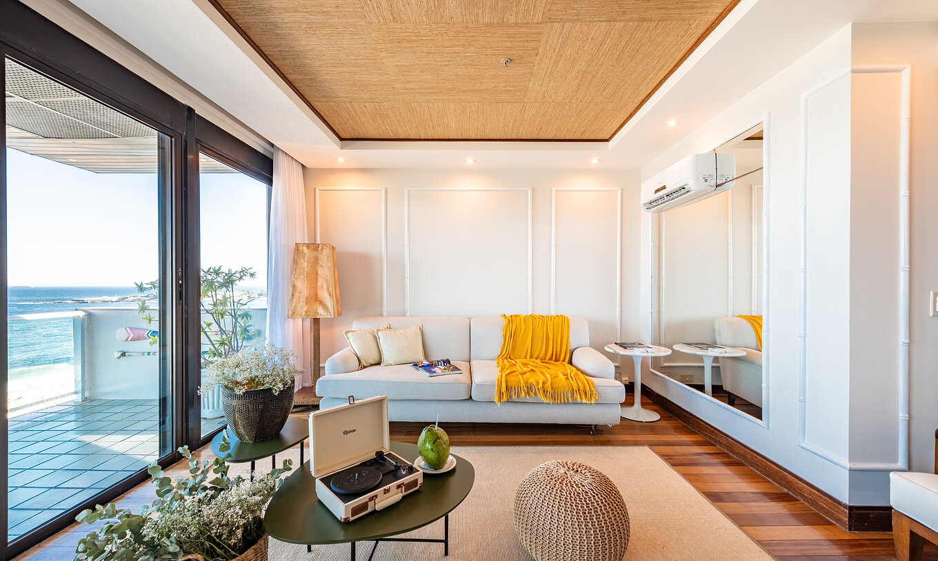 A Suite Presidencial do Pestana Rio Atlântica tem uma sala com teto em madeira, sofá branco e vista para o mar