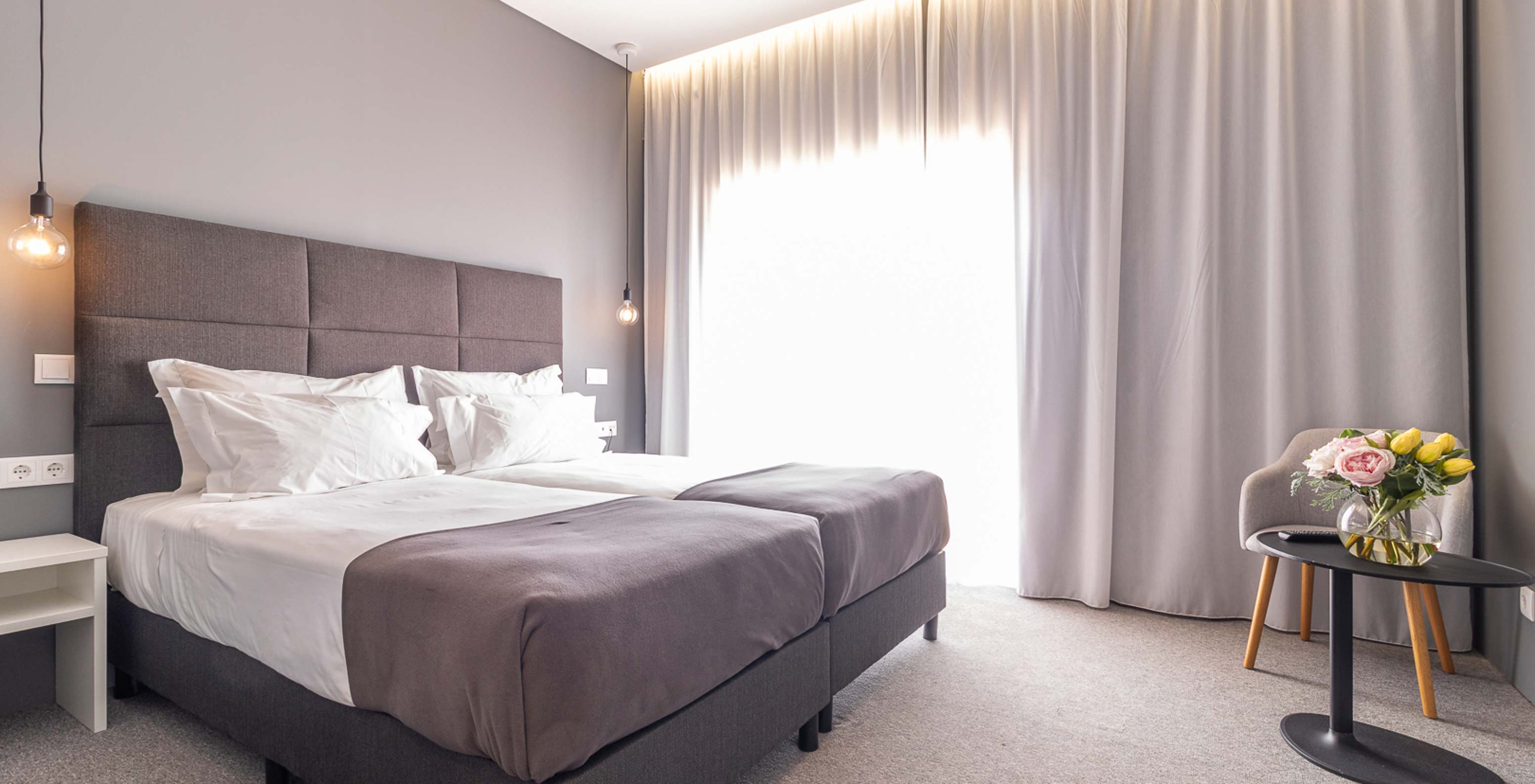 O Quarto Clássico do Pestana Gramacho Residences é um quarto moderno com duas camas, cortinas cinza e mesa com flores