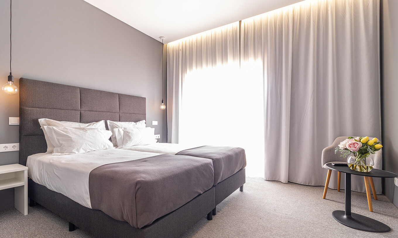 O Quarto Clássico do Pestana Gramacho Residences é um quarto moderno com duas camas, cortinas cinza e mesa com flores
