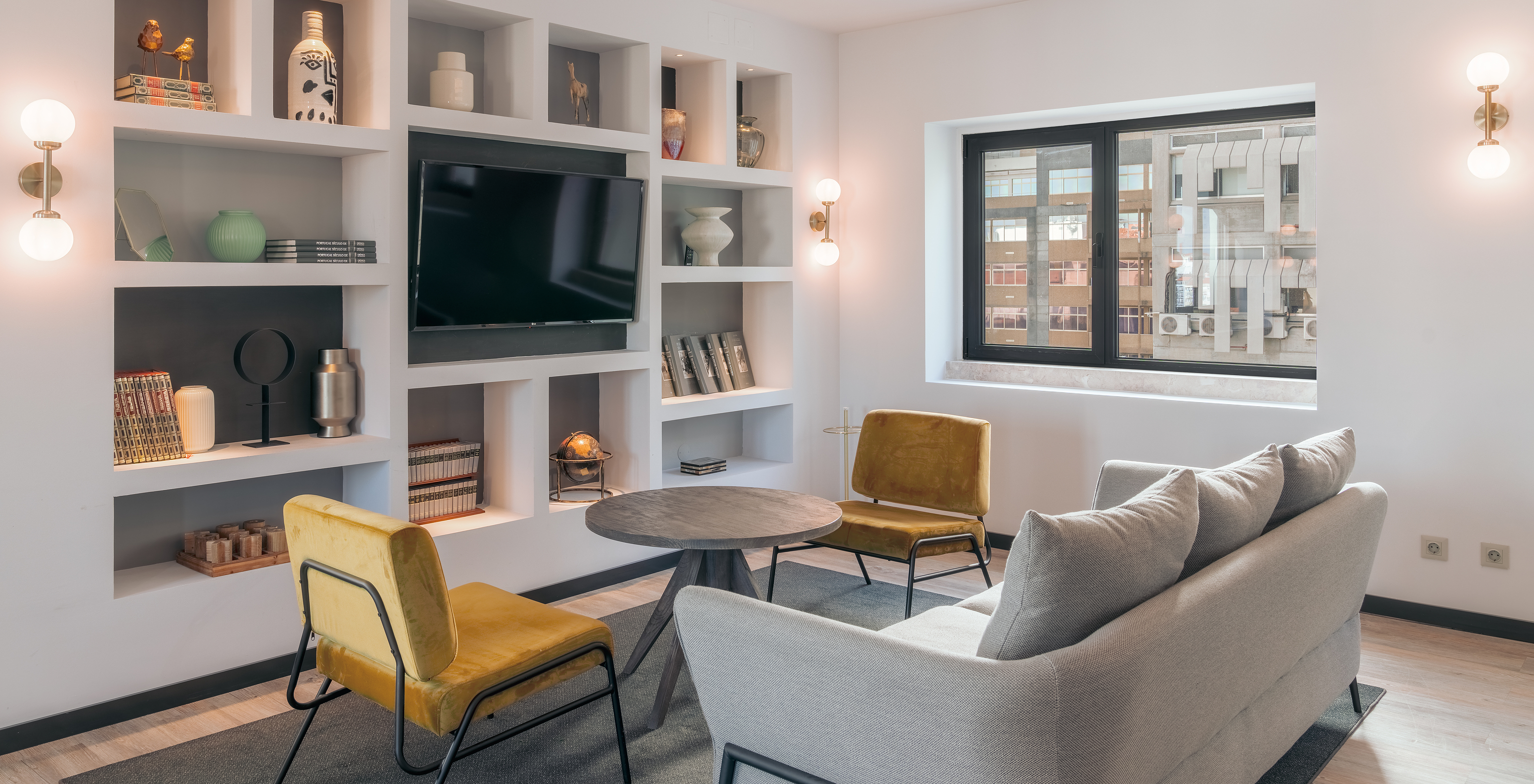 Lounge com sofá, cadeiras, mesa de apoio, televisão, tapete e objetos de decoração com janelas com vista para a cidade