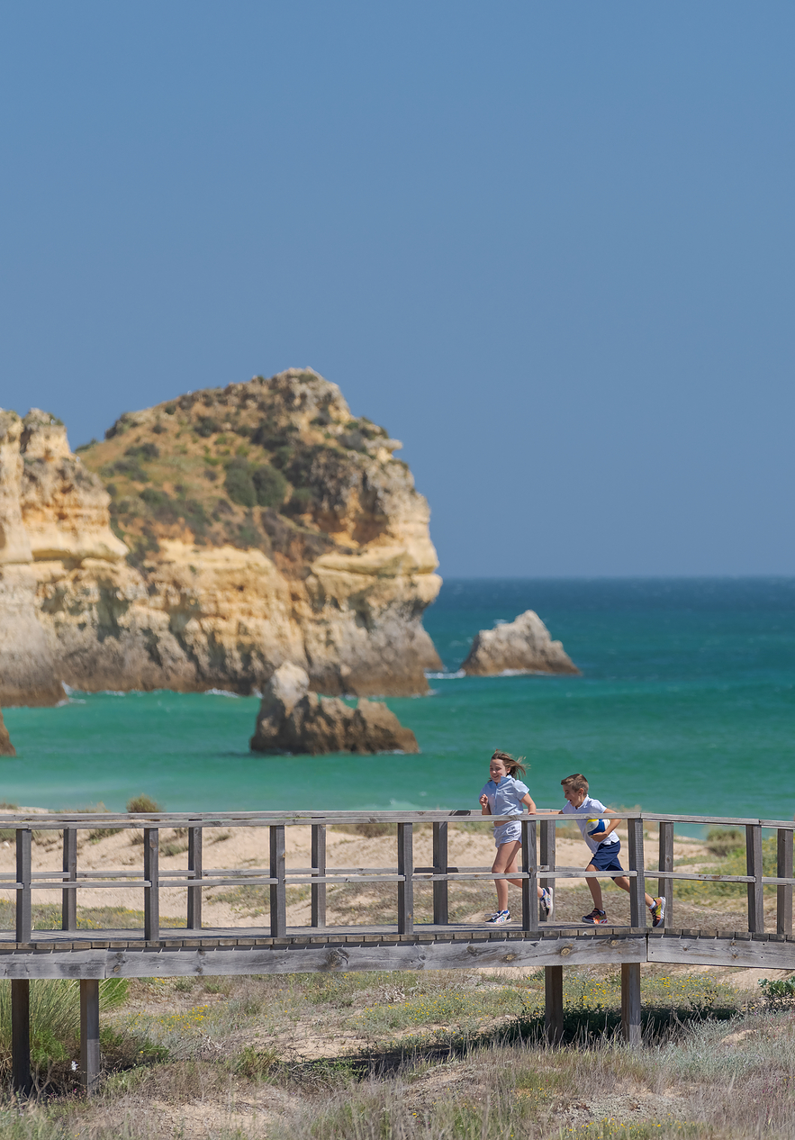 Eltern und zwei Kinder gehen auf einem Holzsteg mit Blick auf das hellblaue Meer und die Klippen im Hintergrund
