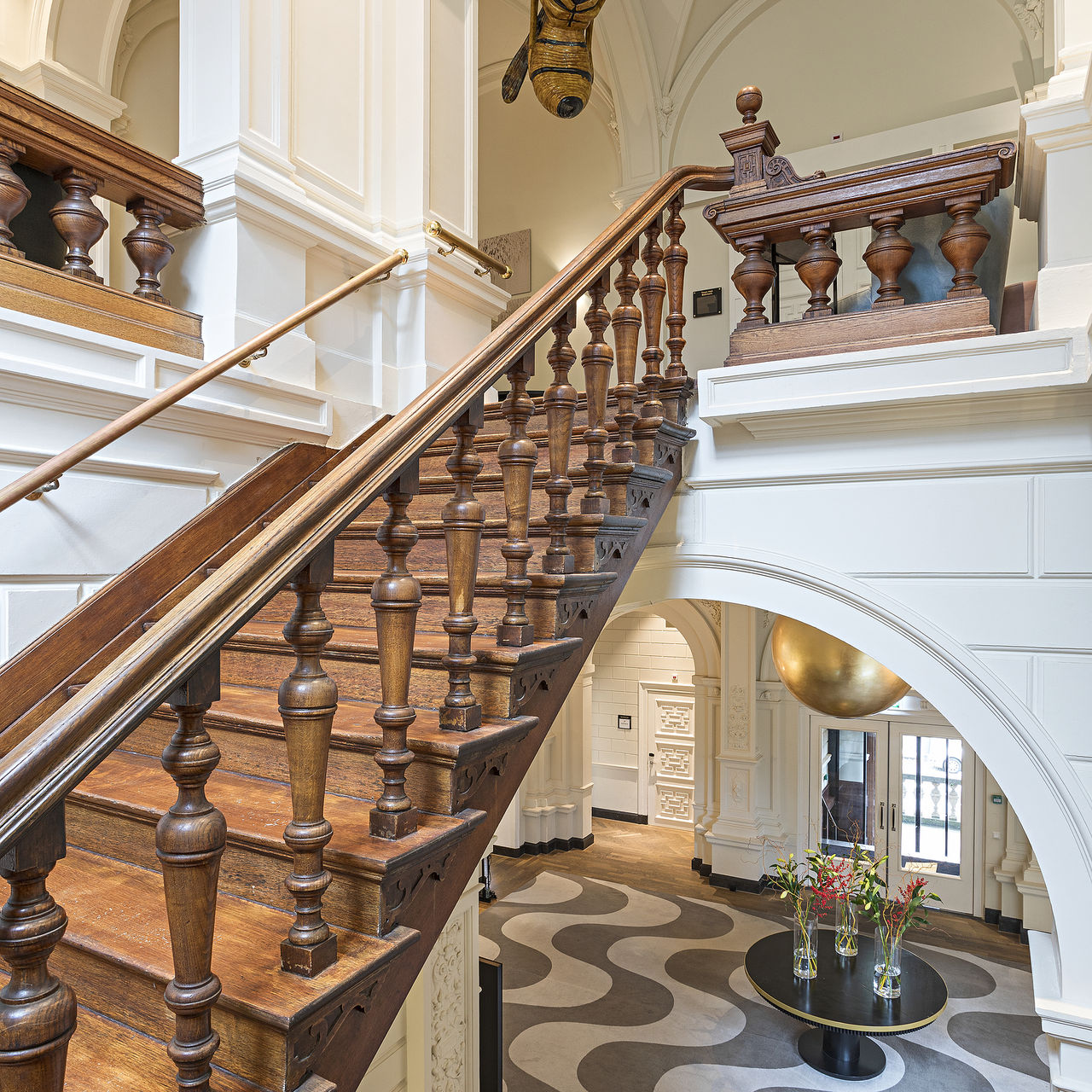 Wooden stairs inside Pestana Amsterdam Riverside, with ornate railing, white walls, and a view to the hall