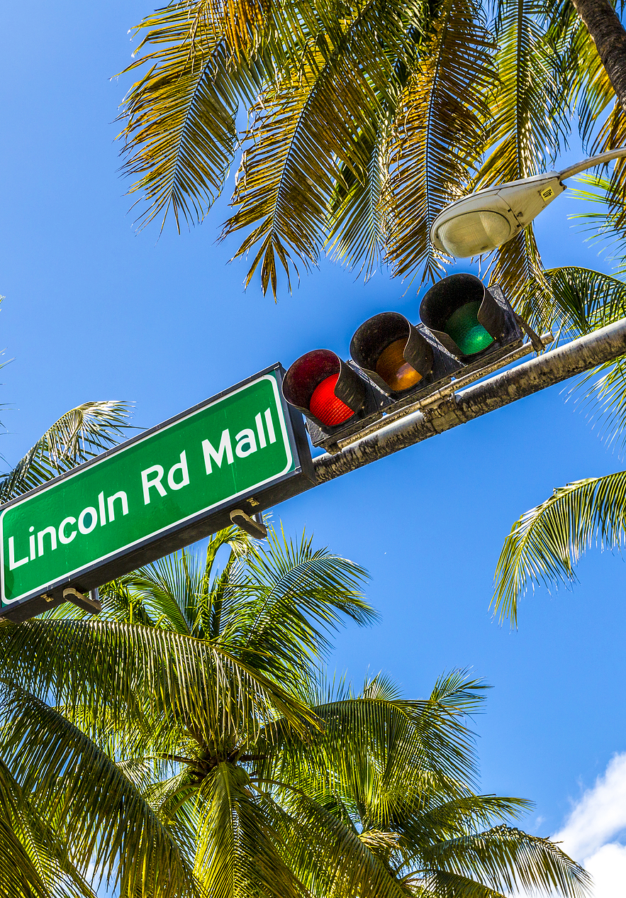 Traffic sign indicating "Lincoln Rd Mall" with a traffic light beside surrounded by palm trees and a clear blue sky