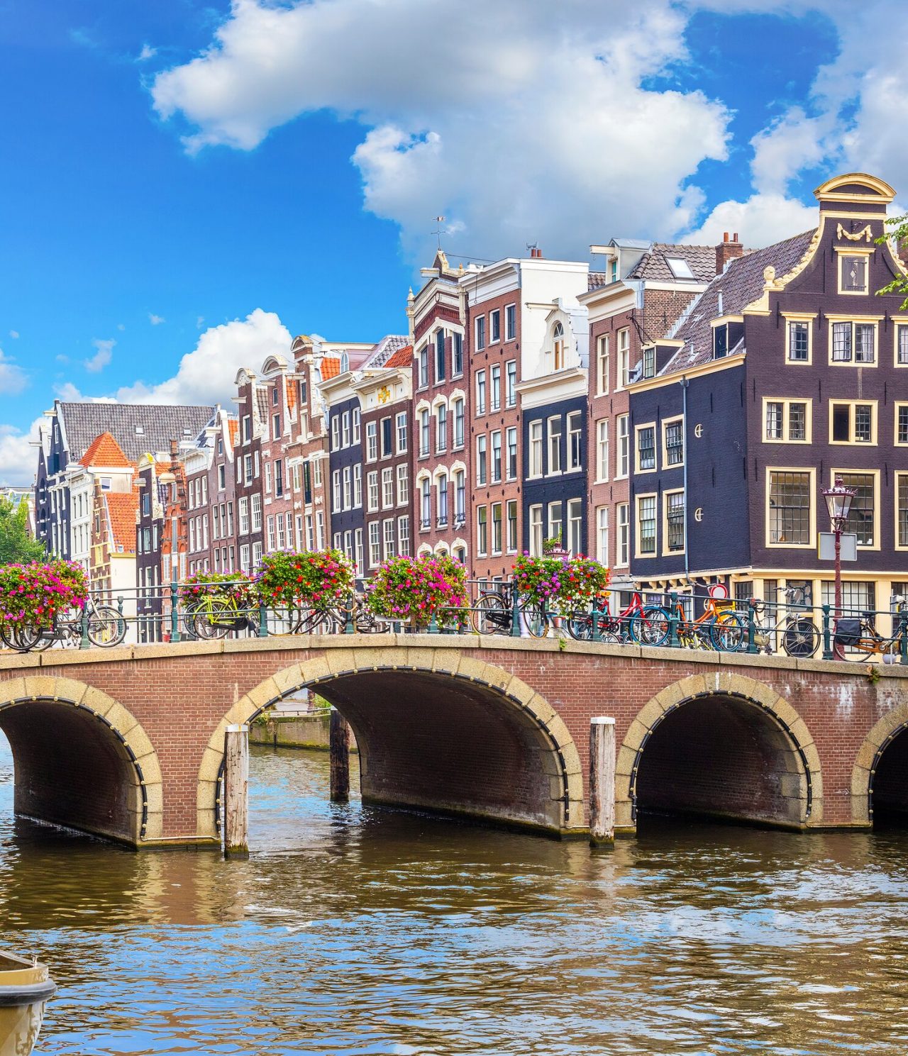 Voetgangersbrug over een kanaal in Amsterdam, met geparkeerde fietsen, kleurrijke bloemen en gebouwen op de achtergrond