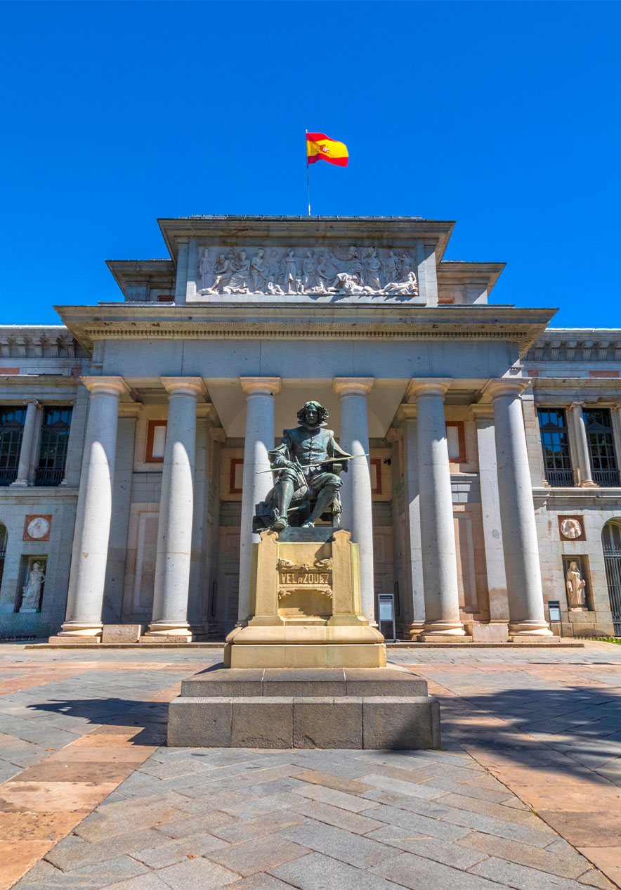 De gevel van het Prado Museum, met een standbeeld bij de ingang en de Spaanse vlag op het dak