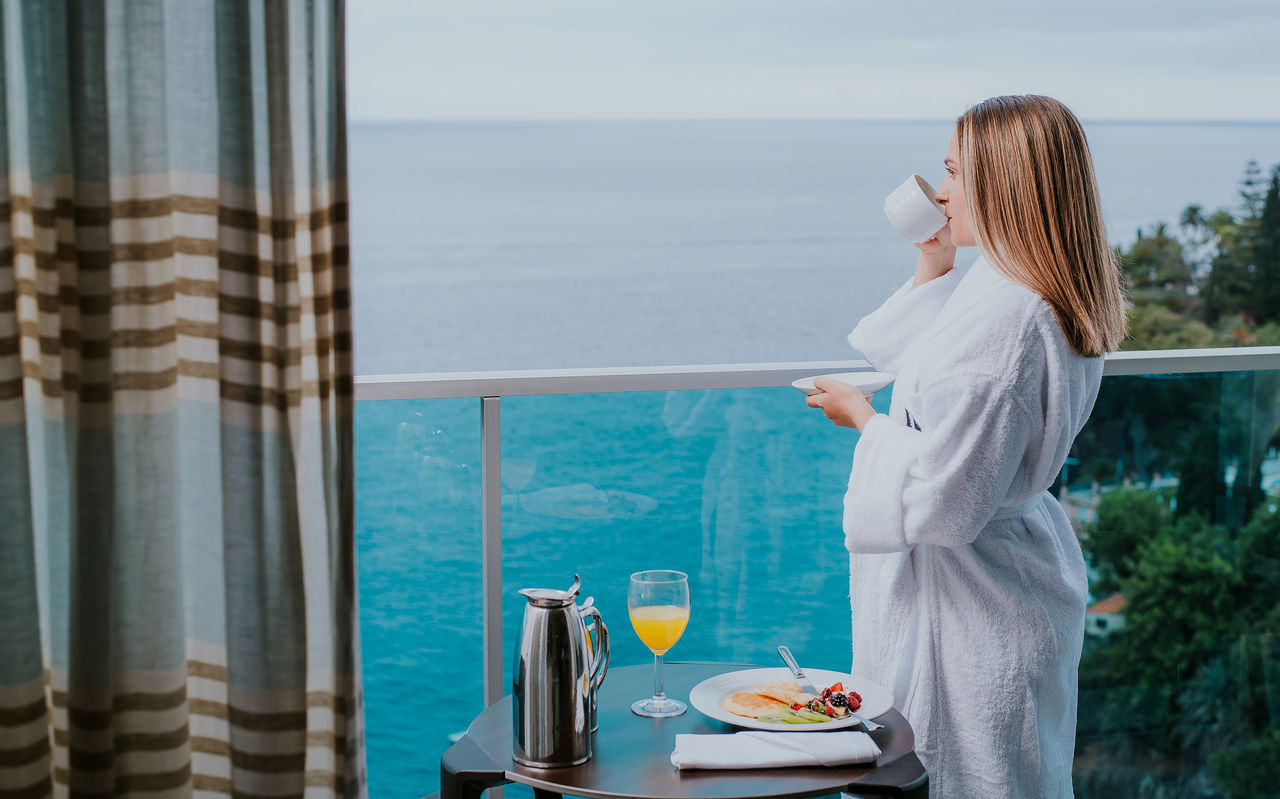 Gast in witte badjas drinkt koffie op balkon met uitzicht op zee, ontbijt en sinaasappelsap op tafel