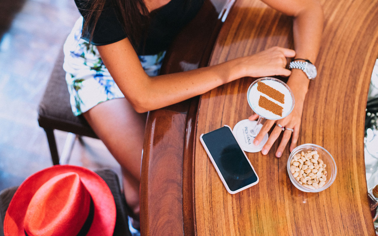 Persoon leunt aan houten tafel met smartphone, drankje met kaneel en schaaltje noten in zonnige setting