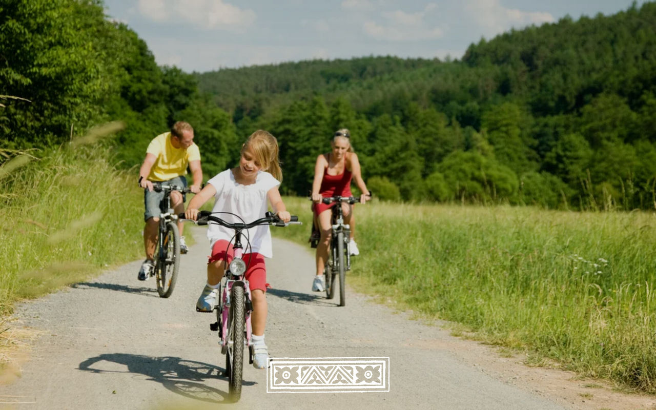 Gezin fietst over een landweg tussen groene velden en bomen onder een heldere zomerlucht met drie fietsers op een zonnig pad