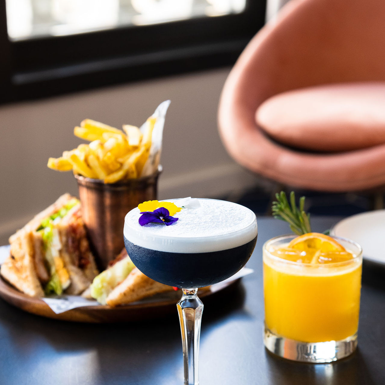 Vista lateral de um cocktail decorado com pétalas, uma bebida de laranja com ervas aromáticas e uma tosta com batatas fritas