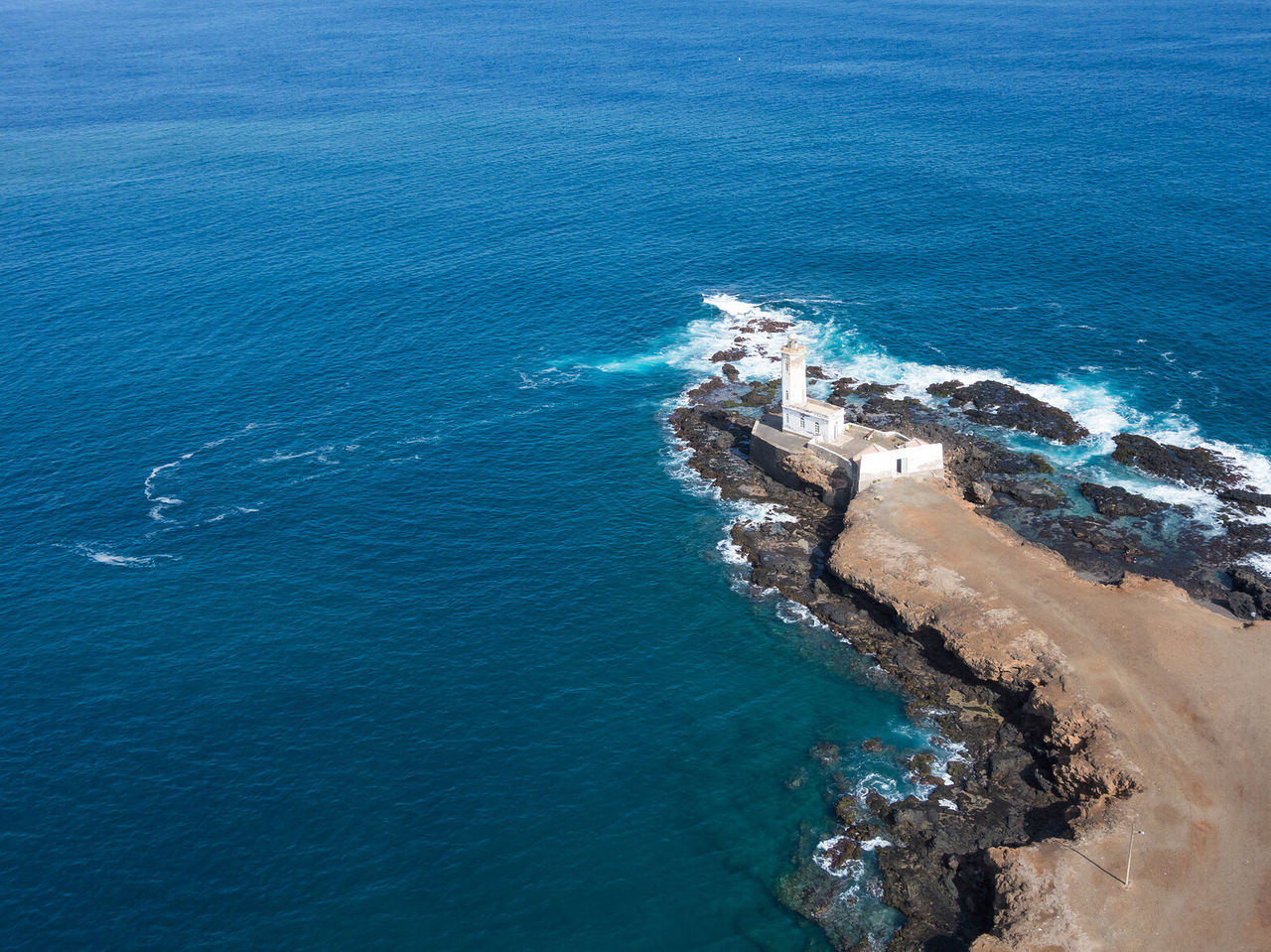 Aerial view of a pier with a lighthouse surrounded by rocks and turquoise blue sea