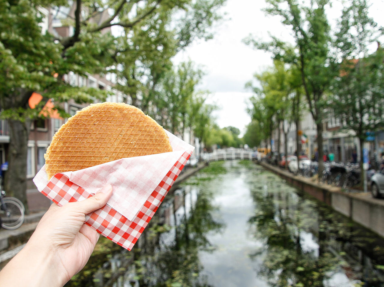Explore Hotels in Amsterdam featuring a hand holding a waffle beside a tree-lined canal and bridge
