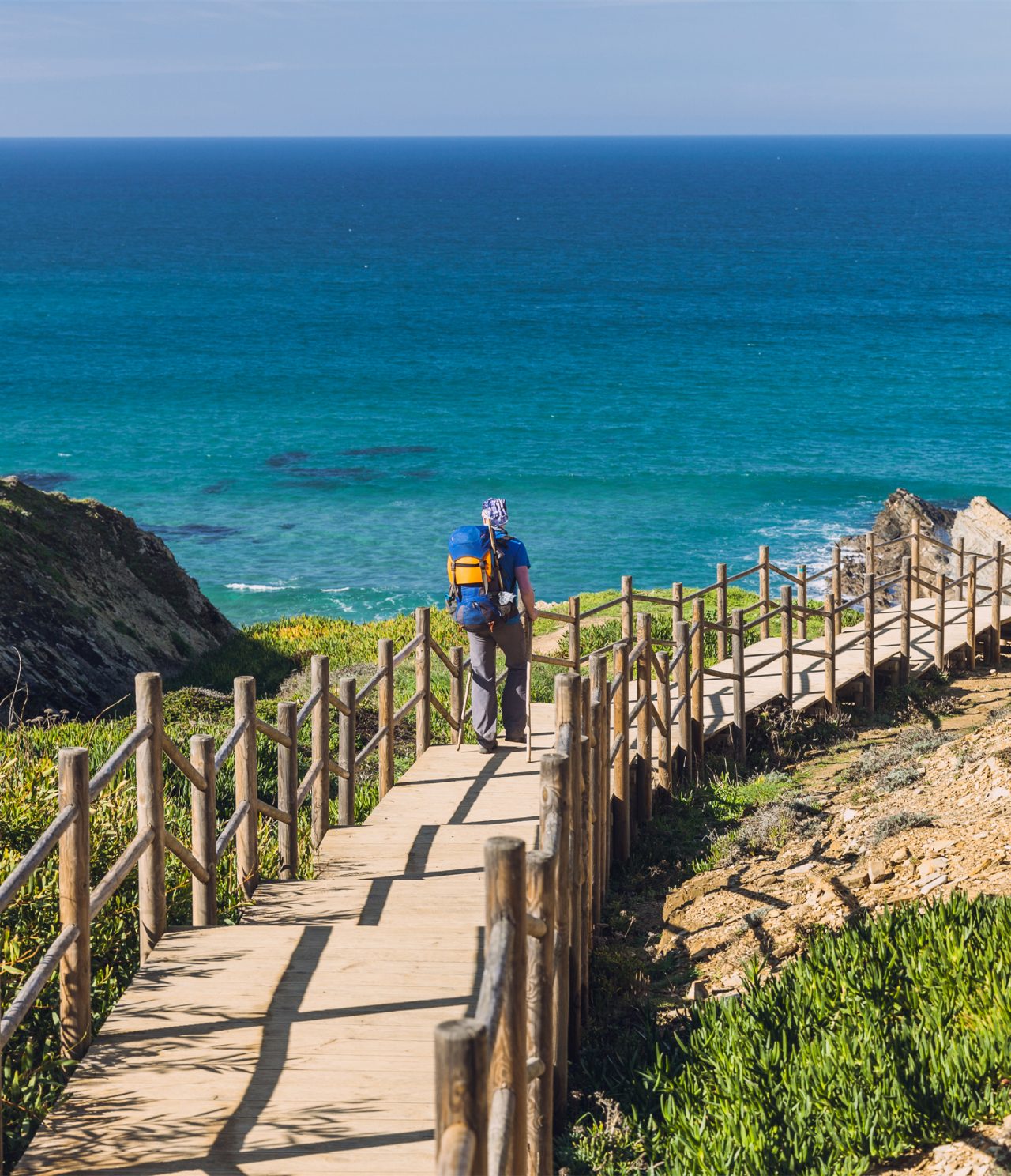 Man walking along a wooden walkway, heading to a beach in Alentejo, with crystal-clear waters