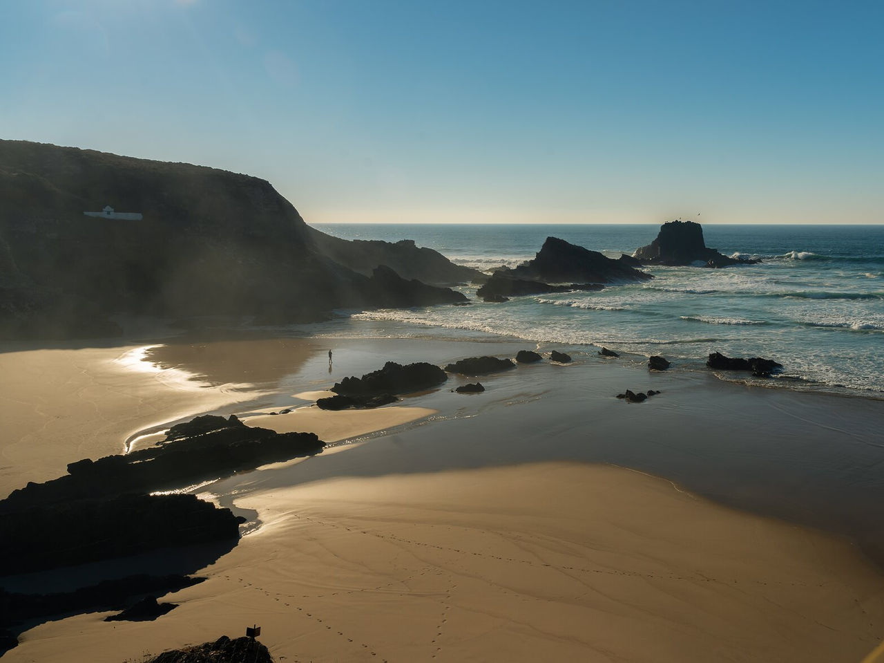 Aerial view of a deserted beach with rocky cliffs, rough sea, and a person walking under a blue sky