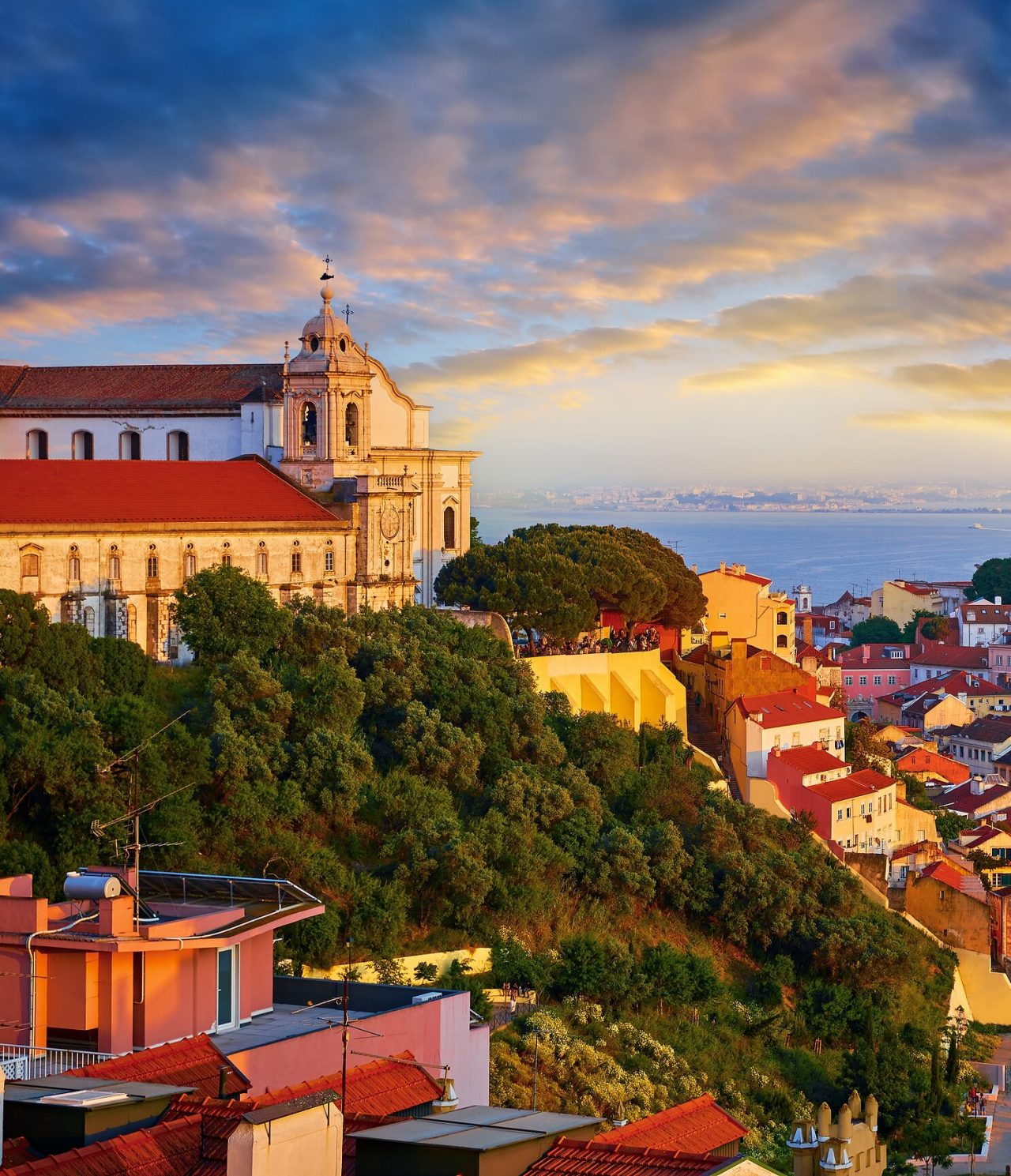 Aerial view of a hill in Lisbon, with a church at the top, the river in the background, and the south bank of the Tagus