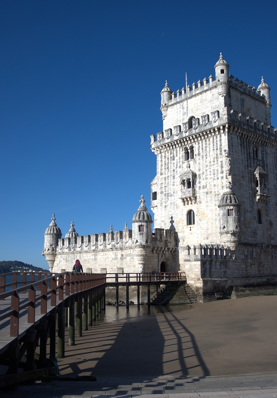 Belém Tower with a wooden boardwalk on the side dark sand and the Tagus River behind the tower with trees around