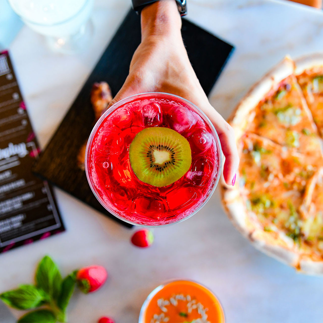 Top view of a pink cocktail with a kiwi slice alongside a pizza meal at Pestana CR7 Funchal
